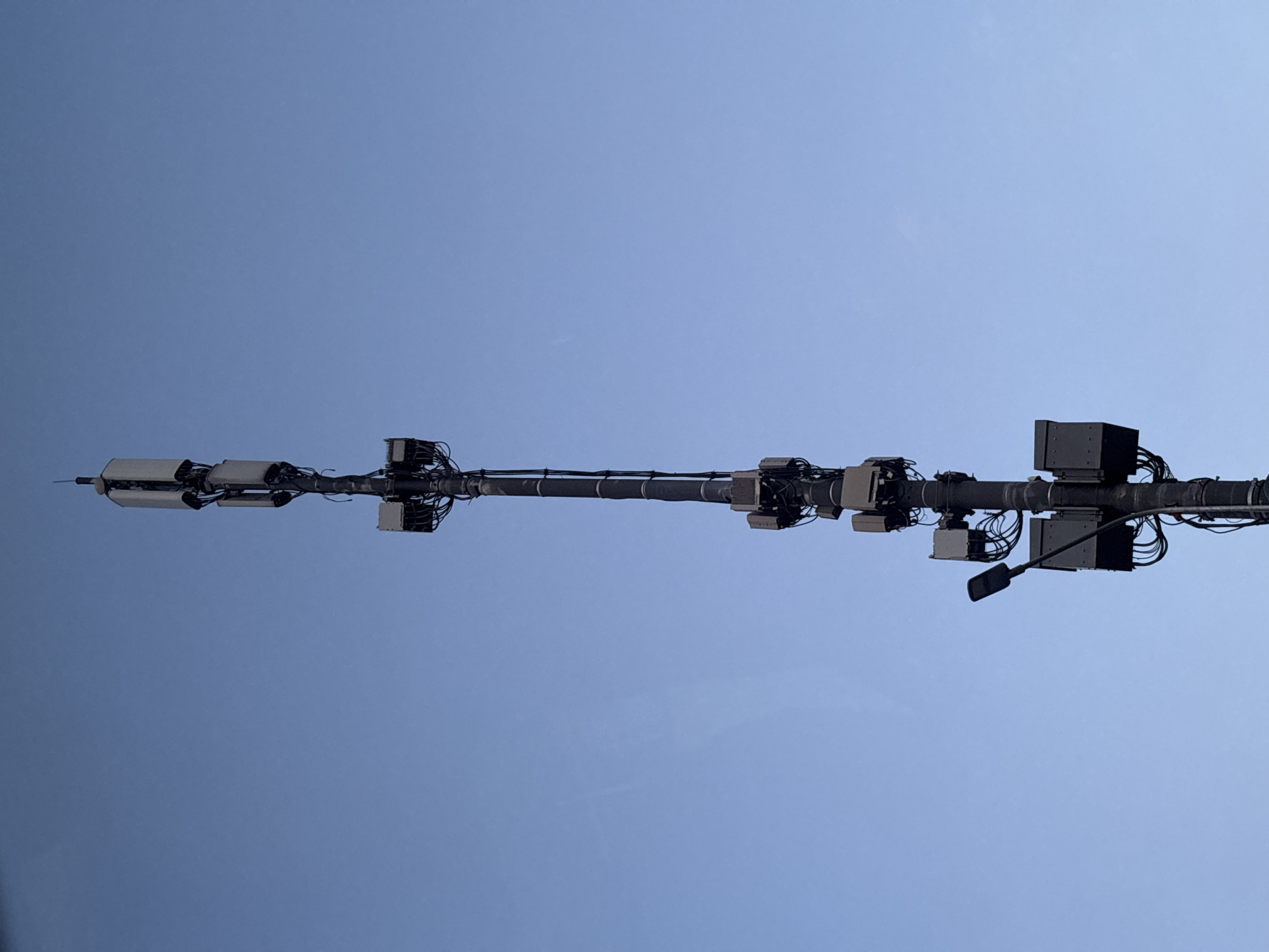 A tall, slender metal pole, likely a telecommunications tower, stands against a clear, pale blue sky. The pole is adorned with various rectangular boxes and antennae mounted at different levels. At the very top, three rectangular panels are stacked vertically, with a thin antenna extending upwards from the uppermost panel. Below these, another set of three similar panels are attached to the pole. Further down, a street light with a dark, angled arm is mounted to the left of the pole, with its light fixture pointing towards the left. Several more rectangular boxes, some with visible wiring bundles, are attached to the pole at various intervals, interspersed with the antennae. The overall scene is stark and functional, emphasizing the industrial nature of the structure against the natural backdrop of the sky.