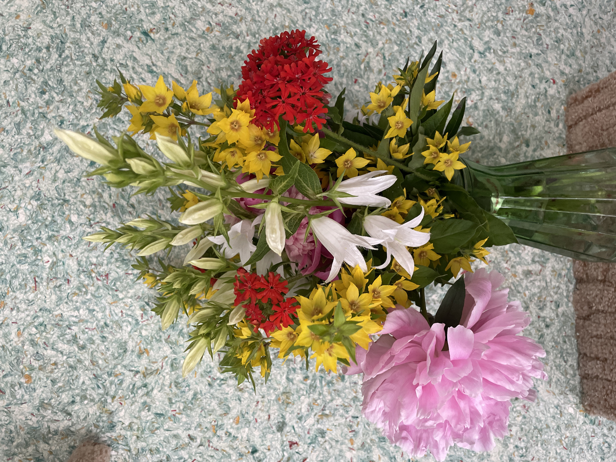 A high-angle medium shot shows a bouquet of flowers in a green glass vase. The bouquet is composed of a large pink peony, white bell-shaped flowers, clusters of small red flowers, and bright yellow star-shaped flowers, all interspersed with green foliage. The vase is placed on a textured surface, possibly a table runner or mat, with a speckled light blue and green wallpaper in the background. The scene is indoors. The location in Perm, Russia is not discernible from the image.