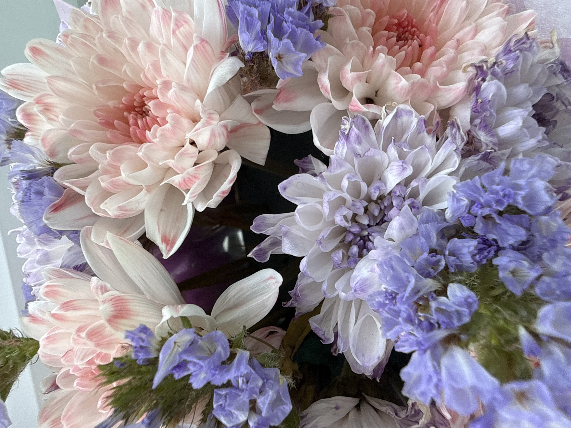 A close-up, overhead shot shows a bouquet of flowers, primarily pink and white chrysanthemums with accents of small purple flowers. The chrysanthemums have layered petals with some tinged with pink at the tips. The purple flowers are delicate and clustered. The background is out of focus and appears to be a soft, neutral color. The overall impression is of a vibrant and delicate floral arrangement.
