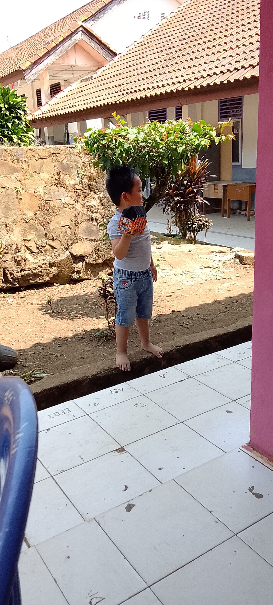 In Jepara, Indonesia, a young boy stands barefoot on a raised dirt border, holding a toy truck. He is wearing a grey t-shirt and denim shorts. Behind him, a stone wall and lush green foliage are visible, with a building featuring a terracotta-tiled roof in the background. The foreground shows white tiled flooring. The scene appears to be outdoors, possibly in a residential area or schoolyard.