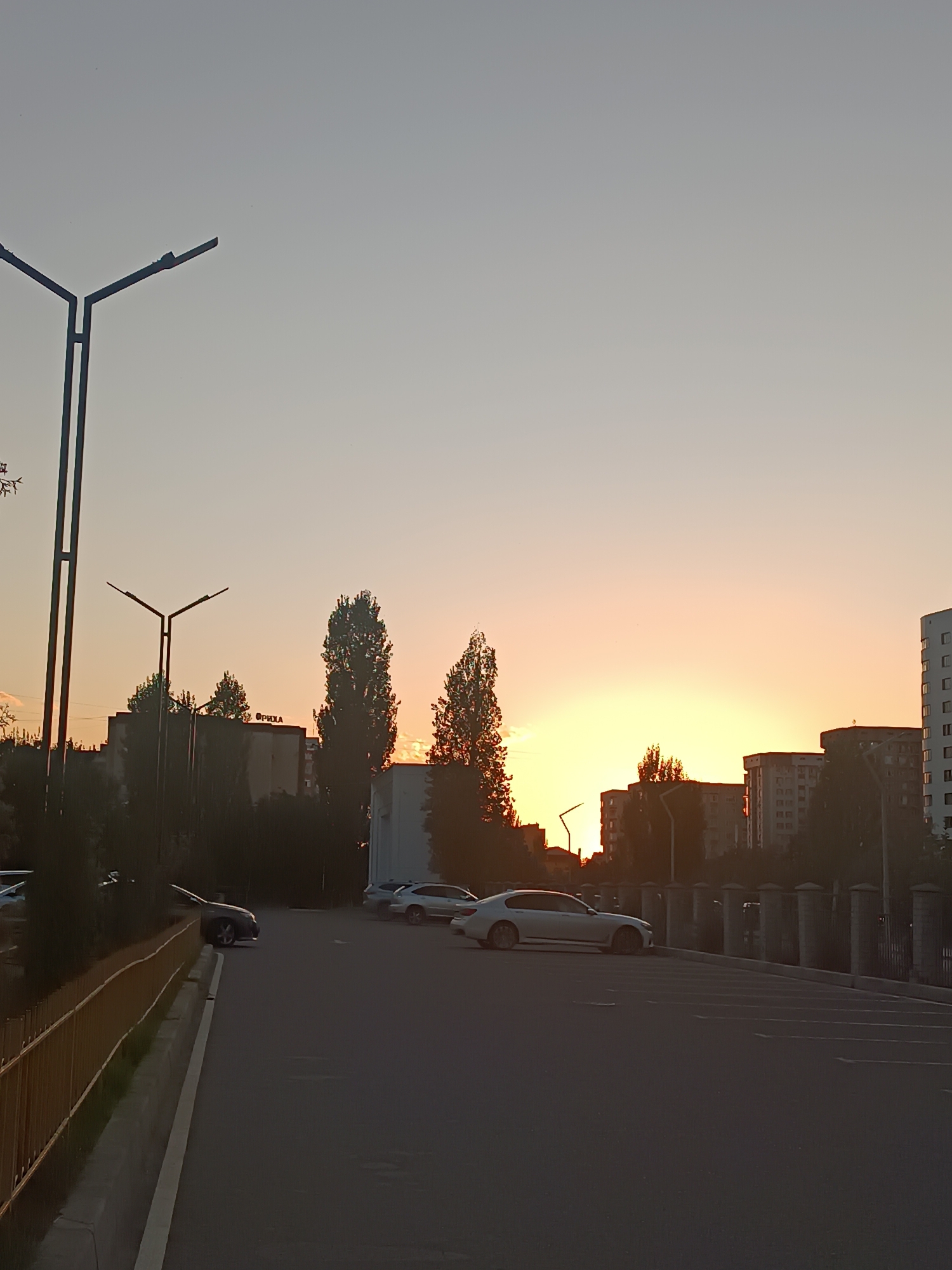 The image depicts a sunset over a city street in Bishkek, Kyrgyzstan. In the foreground, a dark asphalt parking lot is visible, with several cars parked. To the left, a yellow fence runs alongside the street, and a lamppost stands tall. Behind the fence, trees and residential buildings form the urban landscape. The sky is a gradient of pale grey and soft orange, indicating the setting sun. The overall scene is one of urban tranquility at dusk.