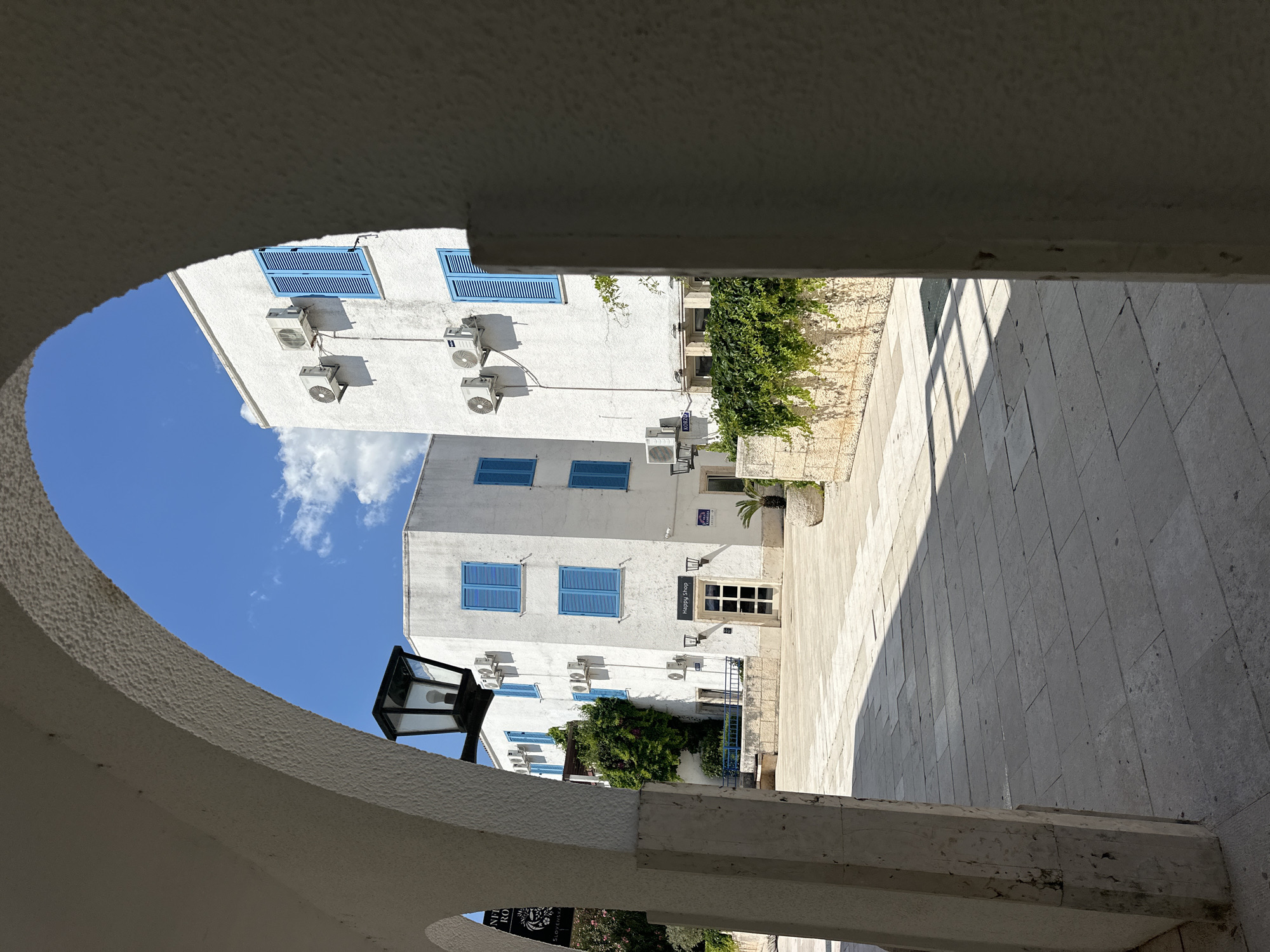 View of white buildings with blue shutters and multiple air conditioning units attached to the facade. Buildings are observed through an archway of a structure, partially seen at the bottom of the image. The buildings appear to be in a sunny location, with a tiled stone path in front of them. It's a sunny day, with clear blue skies showing through the archway. The scene is situated in the city of Budva, Montenegro. The street visible appears to be a pedestrian zone.