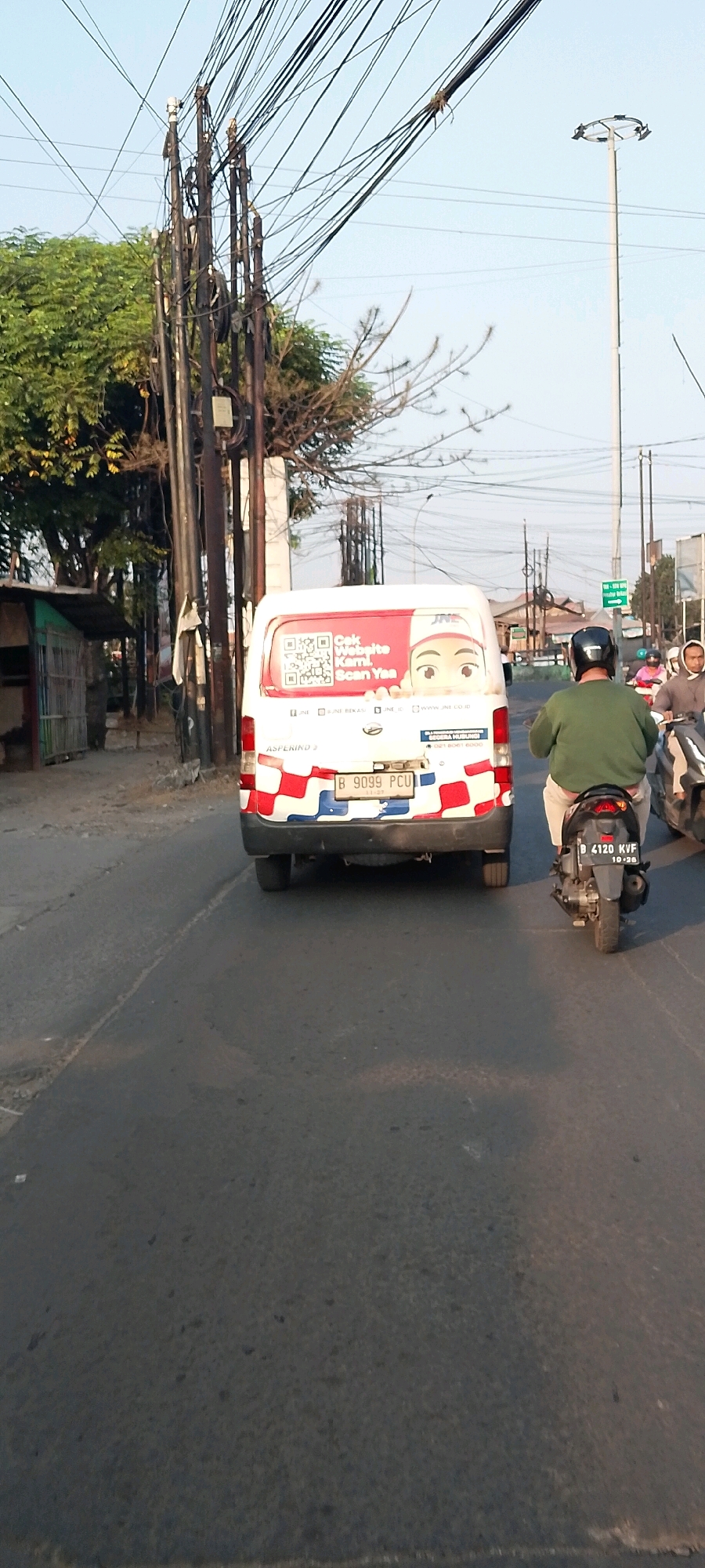 A street-level, rear-view, full-shot image captures a white van with red and blue checkered patterns on its lower rear, parked on a road in Bekasi, Indonesia. The van displays the JNE logo and a QR code, with text that reads "Cek Website Kami. Scan Yaa." In front of the van, a man wearing a green long-sleeved shirt and a dark helmet rides a motorcycle, also with a checkered pattern on its rear. Another person on a motorcycle is partially visible behind him. The road is paved and appears to be in an urban environment with utility poles and wires overhead. Trees are visible on the left side of the image, and a small, simple building with a green roof is also present. The sky is clear. The license plate of the van reads "B 9099 PCU". The license plate of the motorcycle reads "B 4120 KVF".