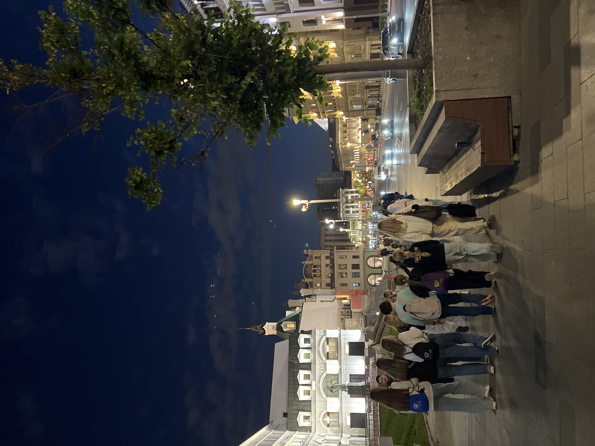 The image captures a nighttime scene on a wide city street, likely in Moscow, Russia, given the architectural style and the visible Cyrillic text on a sign in the background. A group of ten young people, most appearing to be teenagers or young adults, are walking away from the camera on a paved sidewalk. They are dressed in casual, modern clothing, with several carrying backpacks. Some of the backpacks have logos.
To the left of the sidewalk, there is a grassy area and then a building with ornate architecture, featuring multiple arched windows and a domed tower with a spire, illuminated by lights. Further down the street, other buildings are visible, also illuminated at night, suggesting a commercial or residential area.
The street itself is busy with traffic, with cars and vans moving along the road, their headlights and taillights creating streaks of light. Streetlights are also present, casting a warm glow on the scene. A tree stands on the right side of the image, its leaves illuminated by the ambient light, and a wooden bench is situated in front of it. The sky above is dark blue, with scattered clouds and a few distant lights. The overall impression is of a lively urban environment at night.