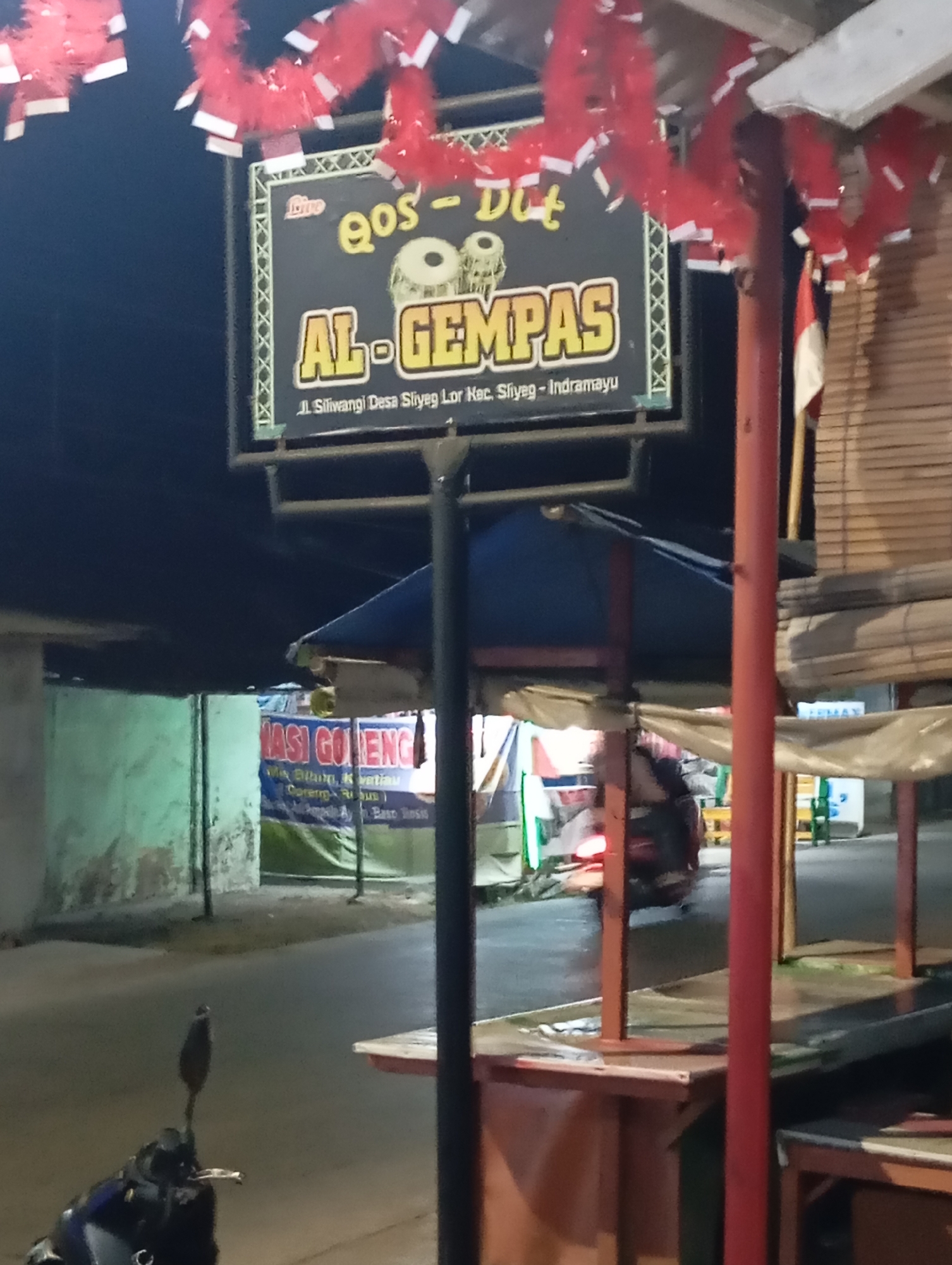 The image shows a street scene at night in Sliyeg, Indonesia. A sign for "AL-GEMPAS" is prominently displayed, featuring graphics of drums and the text "Live Qos-Dut". The sign also indicates the location as "Jl. Siliwangi Desa Sliyeg Kec. Sliyeg - Indramayu". Red and white decorative streamers hang above the sign. In the background, there are various stalls or shops, one with a banner that reads "NASI GORENG". A motorcycle is partially visible in the foreground. The environment appears to be an outdoor market or roadside area with low lighting.