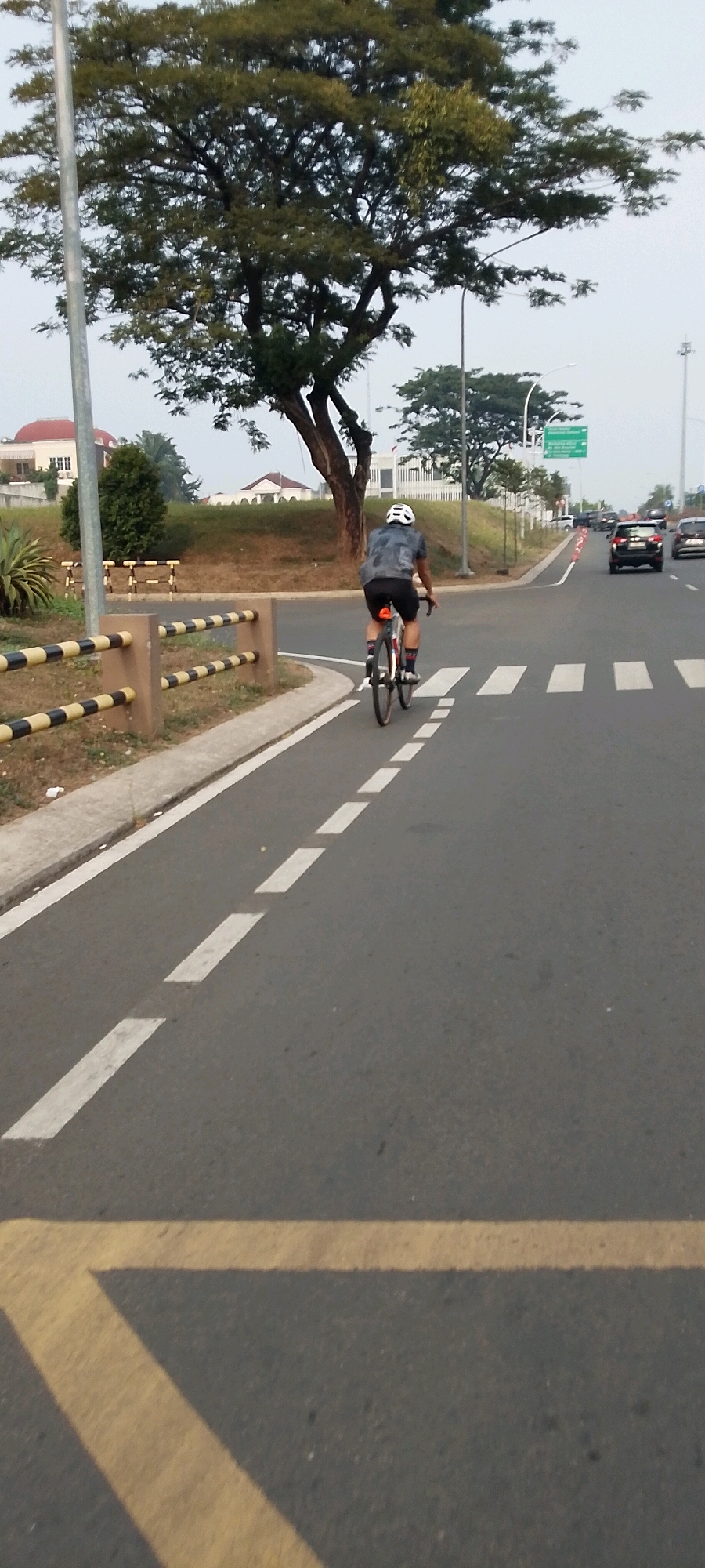 The image shows a cyclist wearing a helmet and a grey and black patterned shirt, riding a bicycle with a black frame and red accents on a paved road. The cyclist is moving away from the camera, on a designated bicycle lane. The road has white dashed lines marking the lane and a pedestrian crossing marked with white stripes. In the background, there are trees, buildings with red roofs, and other vehicles on a multi-lane road. There is also a yellow and black striped barrier on the left side of the frame. The location is identified as Gunung Putri, Indonesia. The sky is overcast.