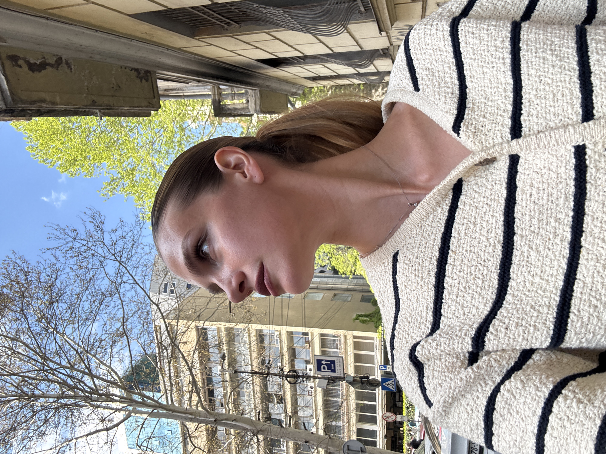 A medium shot, taken outdoors, features the side profile of a person's head, neck, and upper torso. The person, with their hair pulled back into a sleek ponytail, looks downward and slightly to the left with a neutral expression. They wear an off-white, textured knit sweater with distinct vertical dark (black or navy) stripes. A delicate silver-toned chain necklace with a small pendant is visible around their neck.
The background depicts a bright daytime urban scene. Above the person’s head, a clear blue sky with sparse white clouds is visible, along with the vibrant green leaves of a tree, suggesting spring or early summer. Below this, older buildings with multiple windows and balconies line the street. One building has an upper section with intricate, possibly tiled, detailing. In the mid-ground, bare tree branches intermingle with street infrastructure. Several road signs are visible, including a blue square sign with a white "P↑" (indicating parking ahead), another blue "P" sign with an arrow pointing right, and a red-bordered circular sign with a blue background and red diagonal line, typically indicating a 'no stopping' or 'no parking' zone. The overall impression is a candid moment captured on a clear day in a city setting, specifically in Tbilisi, Georgia.