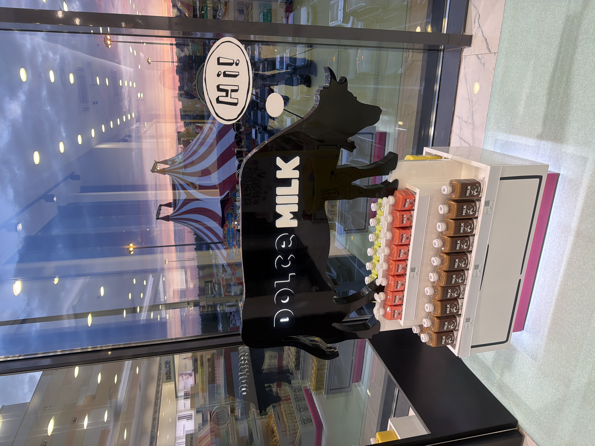 This is an indoor shot of a display for "Dolce Milk" products, likely inside a store or cafe. The scene features a glossy black cow-shaped sign with the words "DOLCE MILK" stacked vertically in white letters. To the right of the sign is a white shelving unit stocked with various flavors of "Dolce Milk" drinks in bottles. The shelving unit has a pink accent color on its side. In the background, there is a large window reflecting the outside environment, showing a view of Novoaleksandrovka, Russia. One can see buildings, streetlights, and a pink sunset sky reflected in the glass. There is also a "Hi!" logo on the window, partially obscured by the cow sign. A striped awning or tent is also visible in the reflection, suggesting an outdoor event or nearby structure. The floor appears to be light-colored, possibly tile or linoleum. The scene is well-lit, possibly by artificial lighting inside the store.