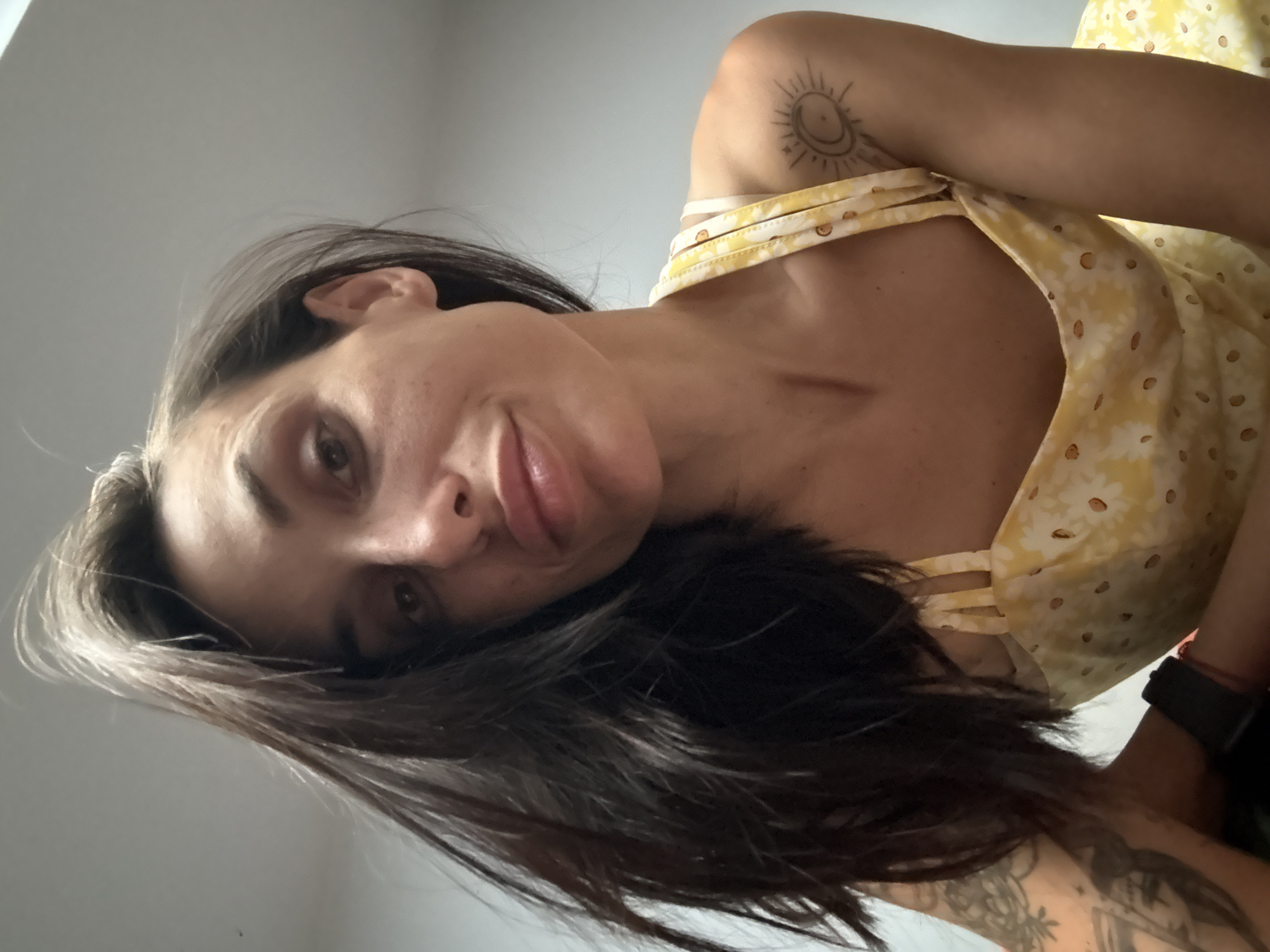 A woman with long, dark hair and a yellow floral dress is captured from a low angle. She is looking at the camera with her mouth slightly open. A tattoo of a sun with a crescent moon is visible on her right shoulder. Her left arm is resting on her chest, and she is wearing a dark watch. The background is a plain, light-colored wall. There are no discernible landmarks or indicators of a specific city or location in the image.