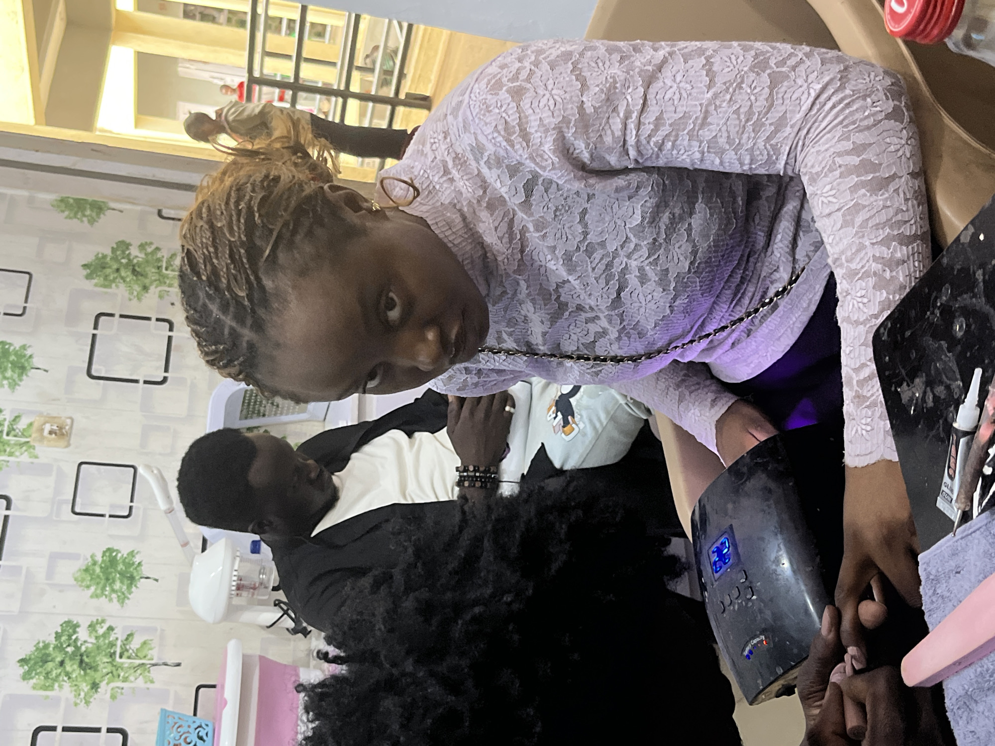 In Jos, Nigeria, a woman with braided hair and wearing a light purple lace top is in the foreground, looking directly at the camera. Her right hand is resting on a black nail dryer which is illuminated by blue LED lights. Her left hand is being attended to by another person whose hands are also visible. In the background, a man wearing a black jacket over a white t-shirt and a beaded bracelet is seated, looking downwards. Behind him, on the wall, is a decorative pattern featuring stylized green trees and black square outlines. Further in the background, through an opening, a person in a light brown jacket and another person in a red top are visible, suggesting an outdoor or semi-outdoor area. The environment appears to be a salon or a similar establishment.