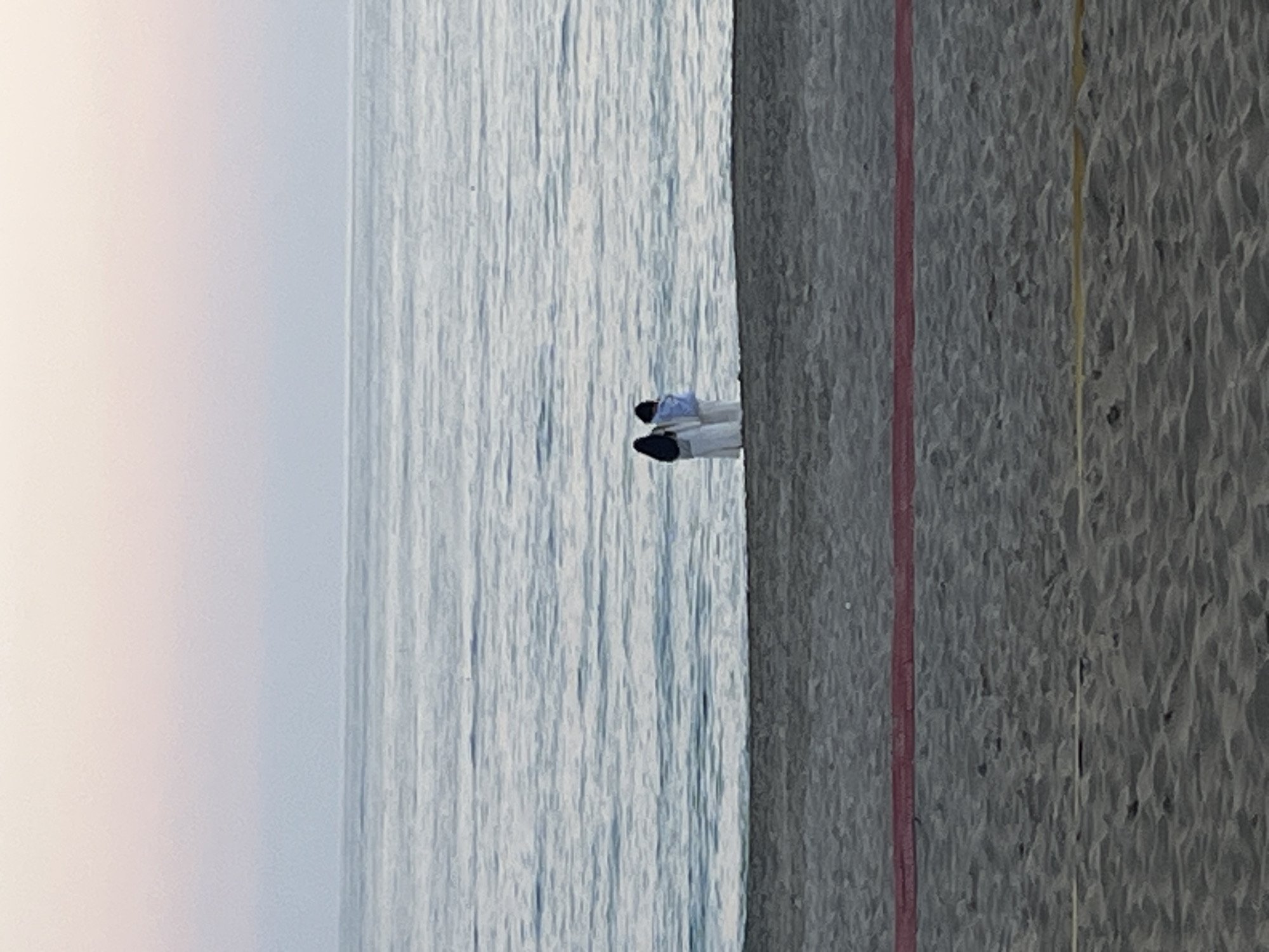 The image depicts a tranquil coastal scene. A vast body of calm water, likely the sea or ocean, occupies the left portion of the frame, extending towards a hazy, light-colored sky at the horizon. The water's surface shows subtle, elongated ripples.
On the right side, a sandy beach is visible, characterized by a textured surface with slight undulations. A single person is situated near the waterline, lying down on the sand. The individual appears to be relaxing or sunbathing, dressed in light-colored clothing, possibly a light blue top and white pants, complemented by dark shoes.
Two thin, distinct lines run parallel to the water's edge on the sandy area. A red line is closer to the water, and a yellow line is positioned further inland from it.
The scene is illuminated by soft, natural light, suggesting either early morning or late afternoon, or possibly an overcast day, as there are no harsh shadows. The overall atmosphere is peaceful and serene. No text is visible within the image.