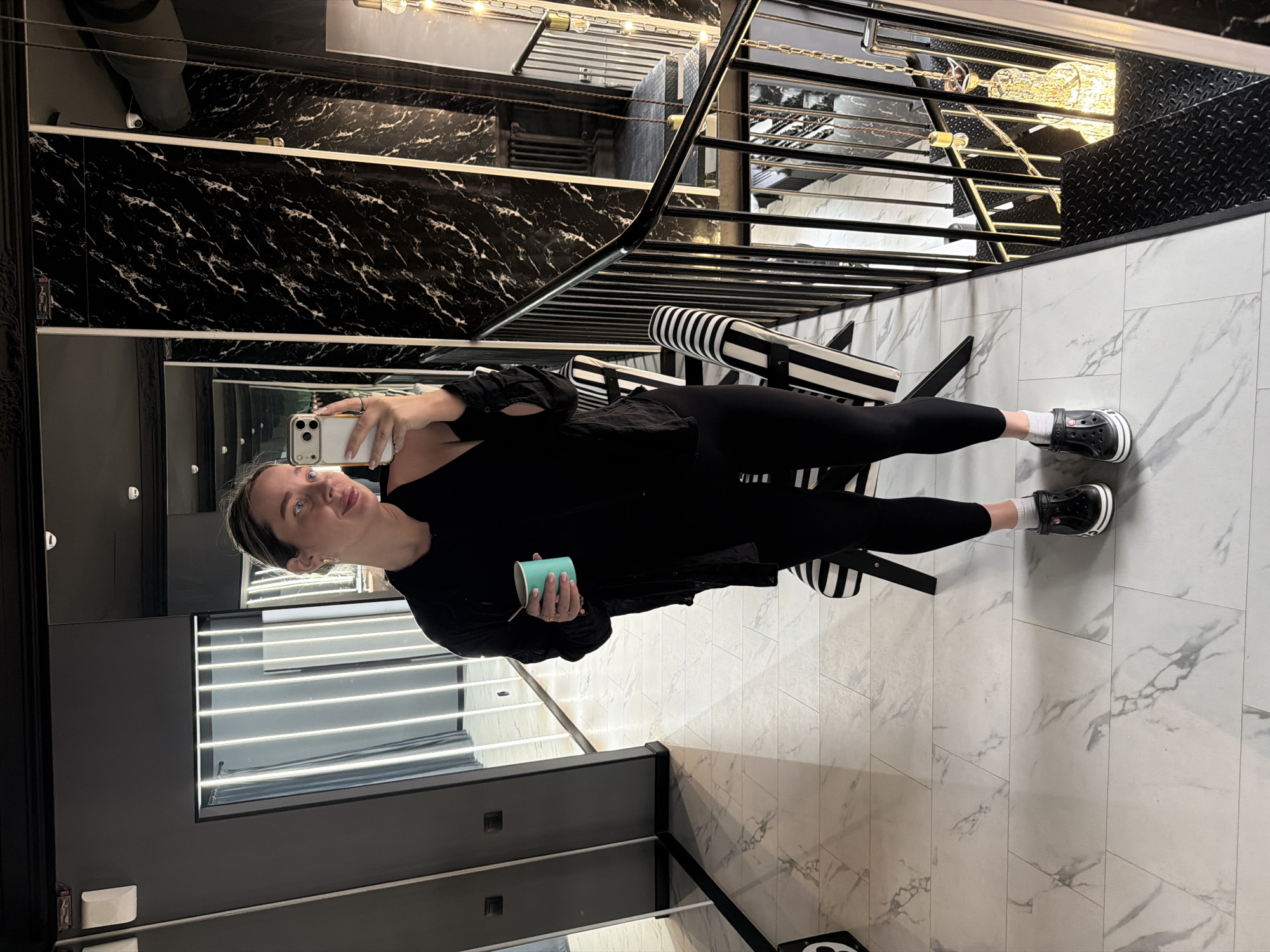 A person is taking a selfie in what appears to be a modern building interior. The person is wearing a black outfit and standing on a marble floor. They are holding a phone in one hand and a small blue cup in the other. Behind them, there is a staircase with black and white striped railings and a decorative lighting fixture on the ceiling. The building appears to be high-end. It is not possible to determine the location in Moscow from the image.