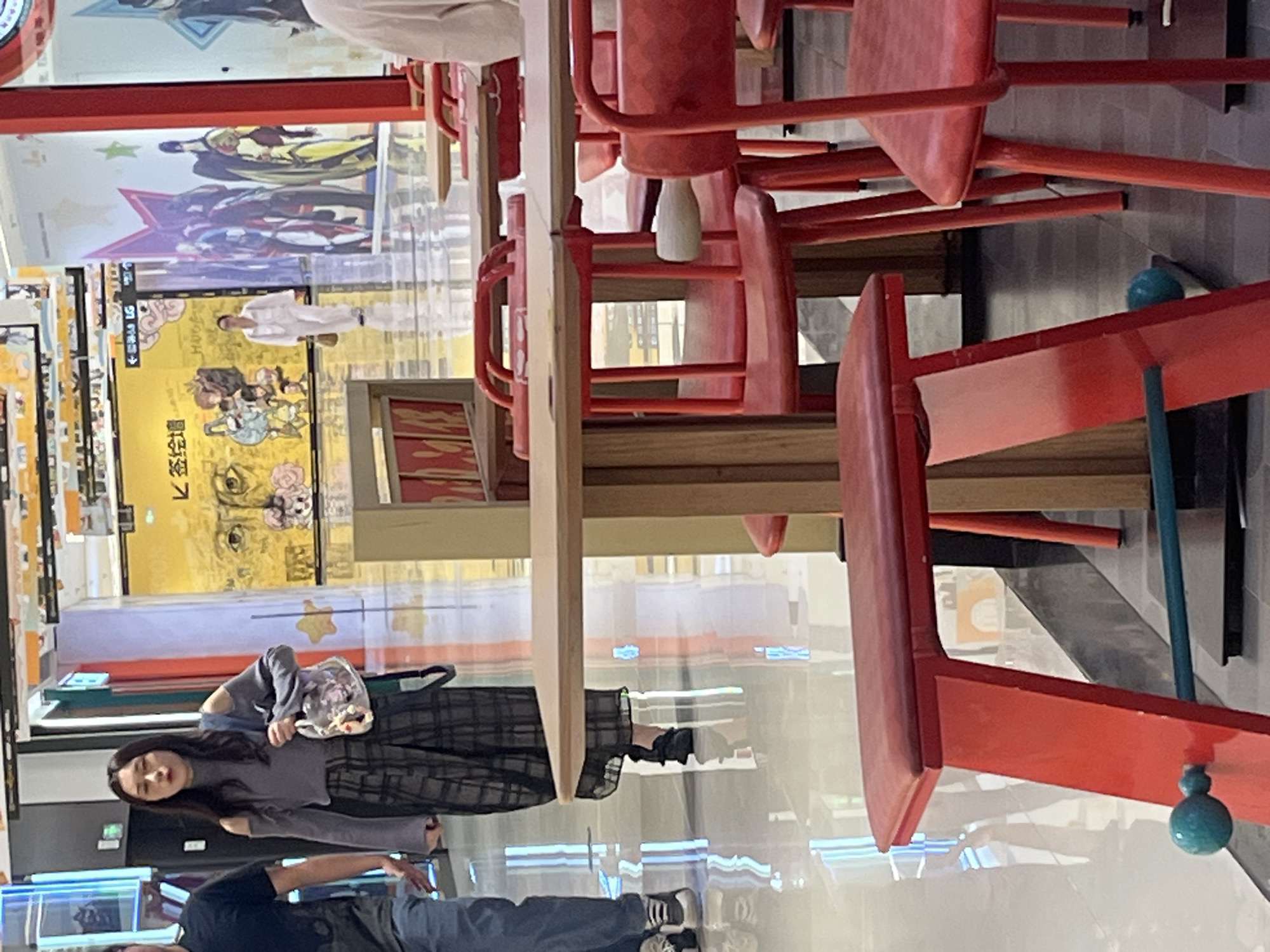 In Yuzhong District, China, a woman is walking through a brightly lit indoor space, likely a mall or commercial area, with a reflective floor. She is wearing a grey off-the-shoulder top with flared sleeves and a black and grey plaid skirt. She carries a translucent bag with colorful charms. In the background, a person in white clothing walks away from the camera, and several vibrant murals and advertisements are visible on the walls. Red chairs and wooden tables are arranged in the foreground, suggesting a dining or seating area.