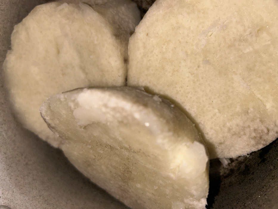Three slices of cooked yam dominate the frame. They are pale, almost white, with a slightly grainy texture. The slices are irregularly shaped and stacked together. The background is a dark, undefined space, possibly a bowl or container holding the yam slices. Close proximity obscures any identifying context.