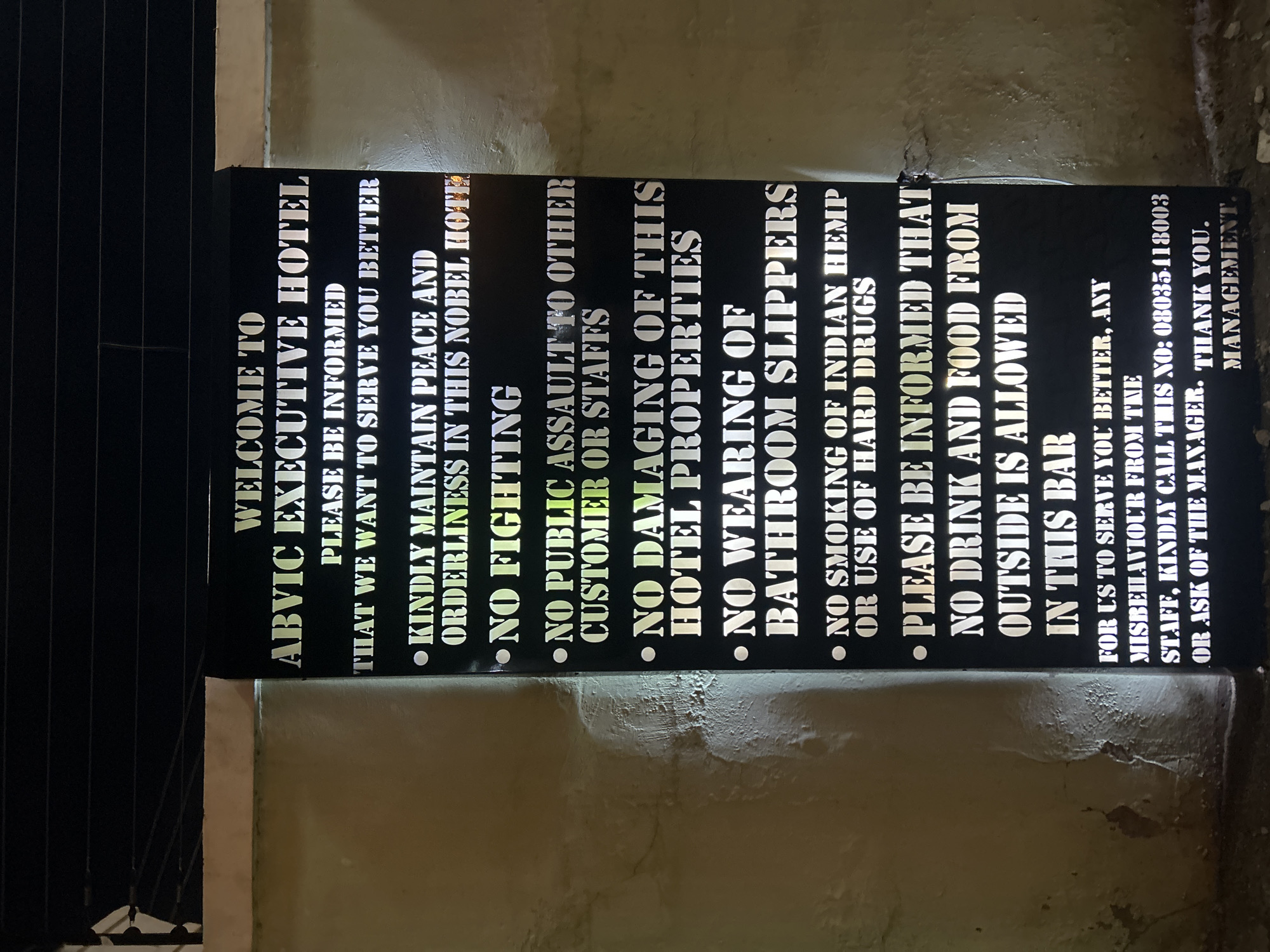 The image shows a sign for the Abvic Executive Hotel, likely placed on an exterior wall of the building. The sign is black with white lettering and lists rules and guidelines for guests. The text includes a welcome message, a request for guests to maintain peace and orderliness, and a series of prohibitions including fighting, public assault, damaging hotel property, wearing bathroom slippers, smoking Indian hemp or using hard drugs, and bringing outside food and drink into the bar. The sign also invites guests to report staff misbehavior to the manager or call a specific phone number. At the end, there is 'Thank you', followed by 'Management'. The environment surrounding the sign appears to be a wall with some texture and possibly discoloration.