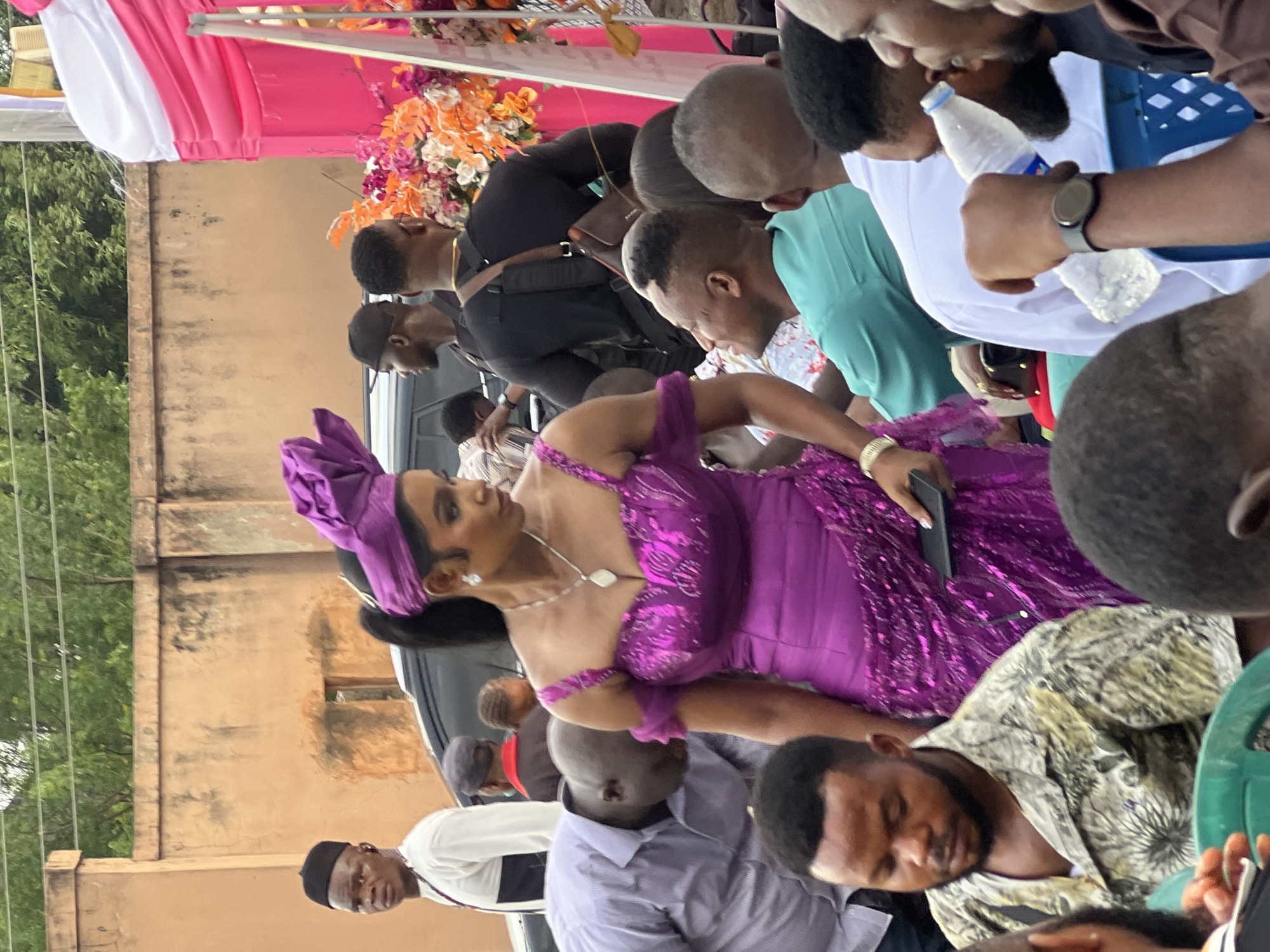 The image shows a group of people, predominantly men, gathered outside, seemingly at a public event. A woman in a bright purple dress and matching headwrap is the focal point, as she is facing to her left while holding a black phone. The people appear to be surrounding her, with some looking in her direction. The background consists of a beige wall and what appears to be a temporary structure decorated with pink and white fabric, as well as colorful flowers. The location appears to be in the city of Unknown, Unknown.