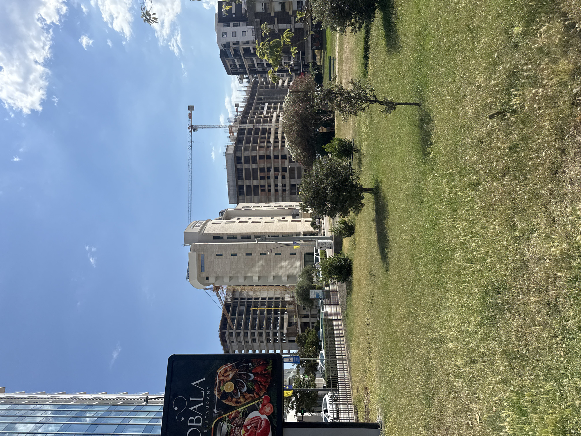 The image depicts an outdoor scene with a focus on urban development and nature. In the foreground, a dark sign for "OBALA RESTAURANT" displays vibrant images of seafood dishes. Behind the sign, a grassy area with scattered trees and a fence runs alongside a street. Several cars are visible parked along the street.
Further back, a large building under construction dominates the midground, with a crane towering over it. To the left of the construction site, another modern building with a glass facade stands. The background features more buildings, some appearing to be residential or office spaces, and a clear blue sky with scattered white clouds.
The overall impression is a blend of urban progress and green space, likely a developed area in Budva, Montenegro. The presence of the construction site suggests ongoing development in the city. The sunny weather and blue sky indicate it is daytime, likely a pleasant day. No people are clearly visible in the image. The scene is static, with no specific activity or event captured, other than the general scene of a city with ongoing construction.