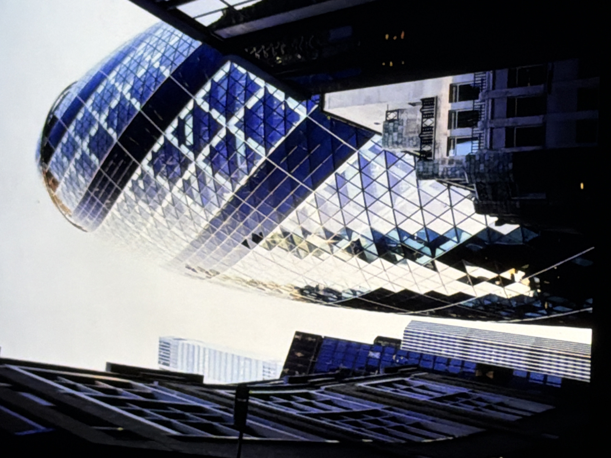 The image presents an upward-looking architectural shot of a modern, ovoid skyscraper with a distinct glass facade, located within the city of Maidstone, United Kingdom. This prominent central building, reminiscent of a gherkin, tapers towards its summit and is constructed from a grid of numerous triangular and diamond-shaped glass panels. These panels display a dynamic range of reflective silvers and deep blues, forming a prominent helical pattern that wraps around the structure. The bright, overcast sky serves as a backdrop, emphasizing the building's unique form.
To the left, a dark, multi-storied building with visible window frames partially obstructs the view, providing a strong contrast against the brighter central structure. Further into the left background, a lighter, tall, rectangular building with vertical and horizontal banding is visible. On the lower right side, an older, more traditionally styled building with stone-like elements, multiple windows, and balconies is partially visible, appearing in shadow. Another dark, undefined building mass is present on the far right. No people, specific actions, or direct interactions are depicted in the scene, which focuses exclusively on the diverse architectural elements within the urban environment.