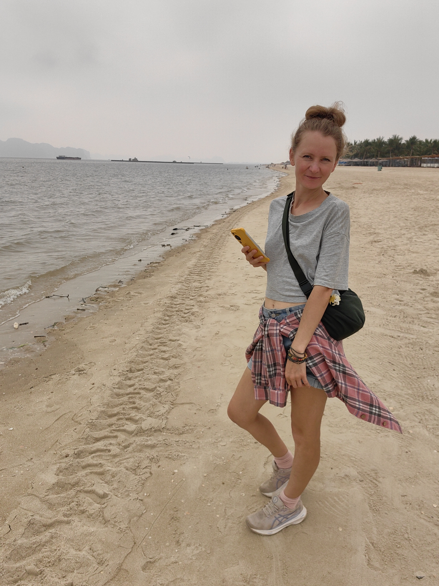 A woman stands on a sandy beach, holding a yellow smartphone. She wears a grey t-shirt, denim shorts, a pink plaid shirt tied around her waist, pink socks, and grey sneakers. A dark green cross-body bag rests on her hip. The beach is sandy, with tire tracks visible. In the background, the ocean meets a hazy sky, with a shoreline visible in the distance. Palm trees and beach umbrellas line a distant portion of the beach. The location is in the city of Bai Chay Ward, Vietnam.