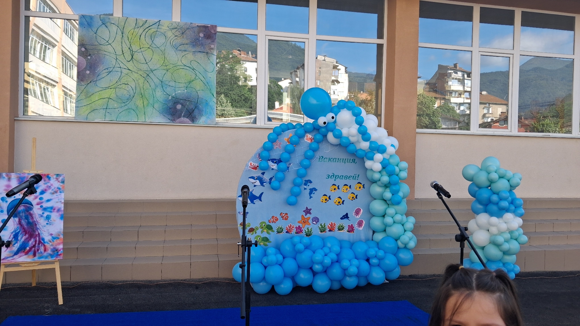 The image depicts an outdoor scene in front of a building, likely a school, in the city of Gotse Delchev, Bulgaria, evidenced by the text "Ваканция, здравей!" (Vacation, hello!) written on a large blue backdrop decorated with underwater imagery and balloons. The backdrop, shaped like a wave or an ocean creature, is adorned with a balloon octopus and various marine life cutouts. To the left of the backdrop, an easel holds a painting of a jellyfish. Microphones on stands are positioned in front of the backdrop and the painting, suggesting a possible event or performance. Two balloon structures, one on the left and another on the right, flank the backdrop. In the foreground, a young girl with pigtails is partially visible, looking towards the camera. The background shows windows reflecting buildings and mountains, indicative of an urban setting.