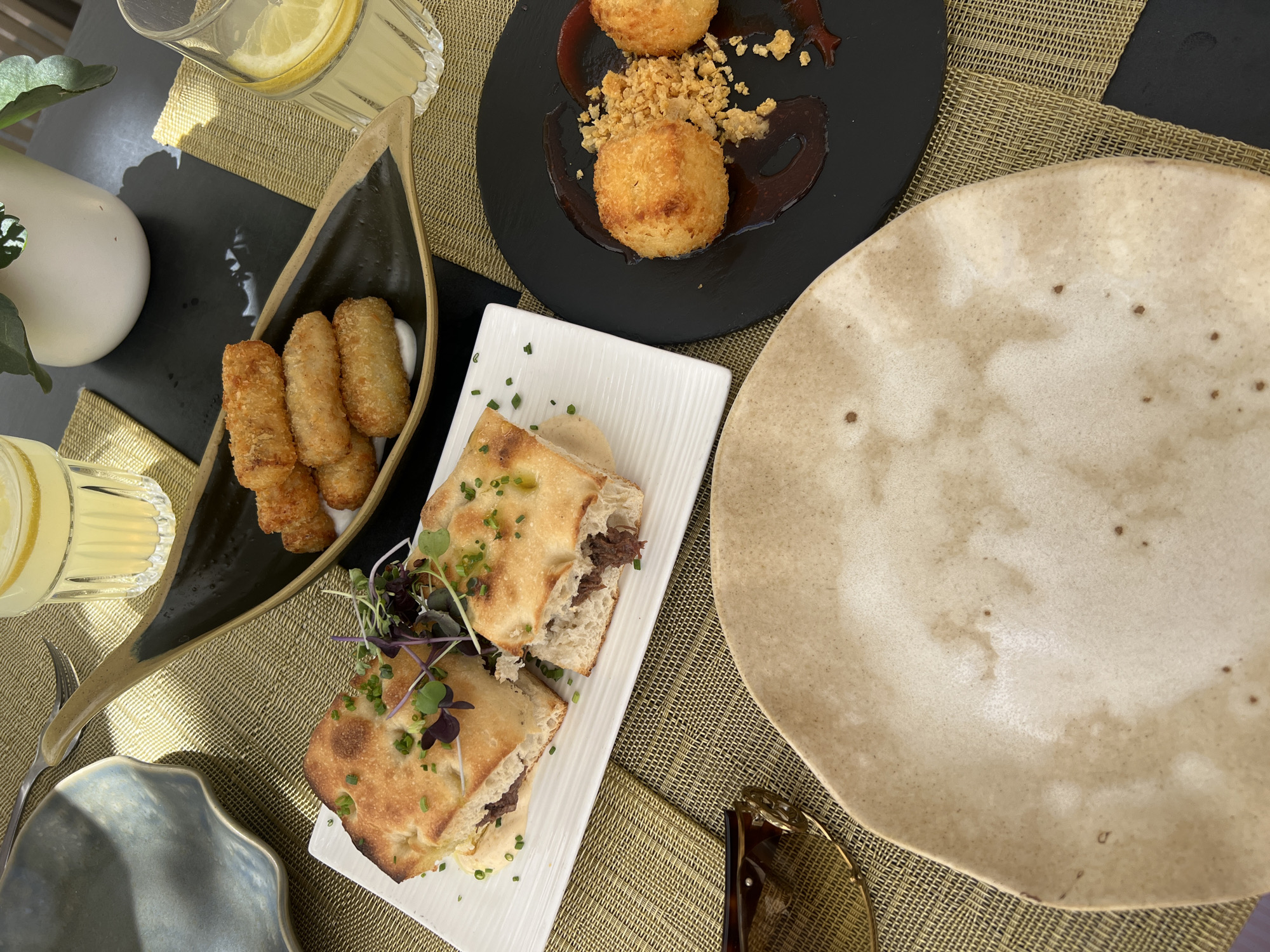 A close-up, overhead shot captures a table setting with various food items and drinks. In the foreground, a beige, irregularly shaped plate is empty, positioned on a woven, gold-colored placemat. To the left of this plate, a pair of tortoiseshell sunglasses rests on the placemat.
Behind the sunglasses and plate, a white rectangular plate holds a halved sandwich on focaccia bread, garnished with chives and microgreens. The sandwich filling appears to be shredded meat. Adjacent to this, a dark, leaf-shaped dish contains several golden-brown, fried croquettes served on a white sauce.
Further to the right, a black circular plate presents two golden-brown, round fritters or arancini, accompanied by a dark sauce and a sprinkle of crumbled breadcrumbs. Two glasses of lemonade, each with a lemon slice, are visible, one behind the croquettes and another partially visible at the top right. The setting is outdoors, suggested by the dappled sunlight and the greenery of a plant in a white pot in the background. This scene is likely set at a restaurant in the charming town of Pollença, Spain, known for its picturesque streets and culinary offerings.