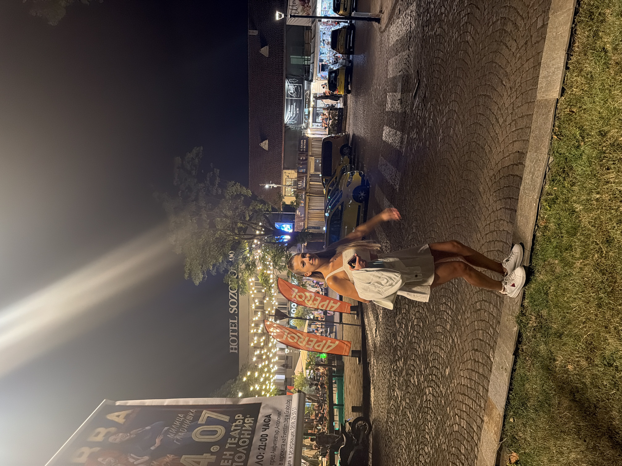 A young woman in a white dress and white sneakers stands on a cobblestone street at night in Sozopol, Bulgaria. She is holding a white jacket and a phone in her hand. The street is lit by string lights above a row of restaurants and shops, and a sign for "HOTEL SOZOPOL" is visible. In the background, there are several cars, including yellow taxis, and two orange flags with the "Aperol" logo. A large banner advertising an event with the date "4.07" and Bulgarian text is on the left. The scene suggests a lively evening in a tourist area.