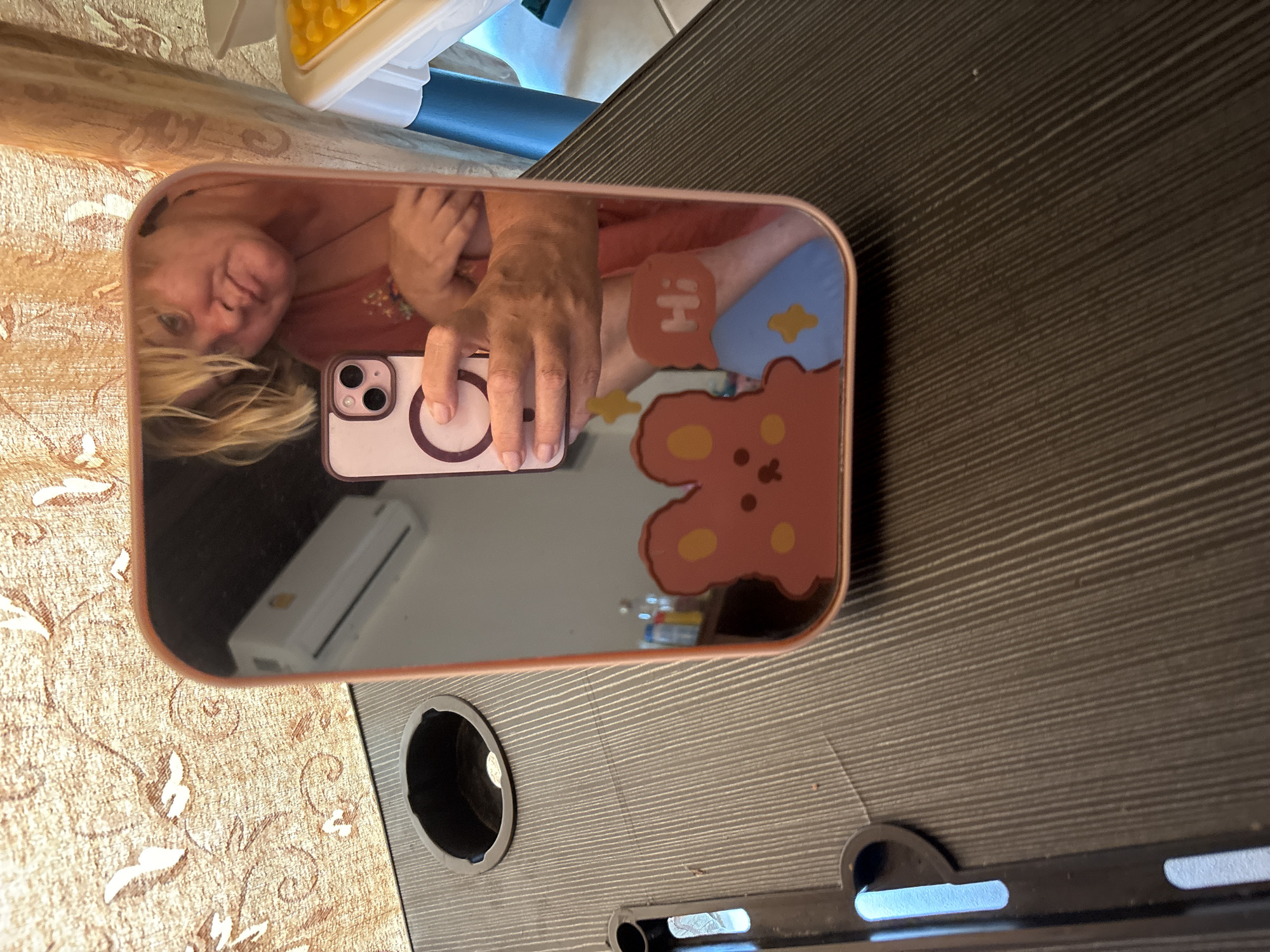 The image shows a mirror reflecting a person taking a picture of themselves with a phone. The reflection includes the person's face, hand holding the phone, and a sticker on the phone case. The surrounding environment in the reflection includes an air conditioning unit and possibly other objects. The mirror is placed on a table with a built-in cup holder and what appears to be a stand. The table has a dark, textured surface. There is a carpet or patterned textile partially visible in the corner of the image. The location appears to be indoors. It is not possible to confirm that this scene is in บ้านบางรักษ์, Thailand, without additional information.