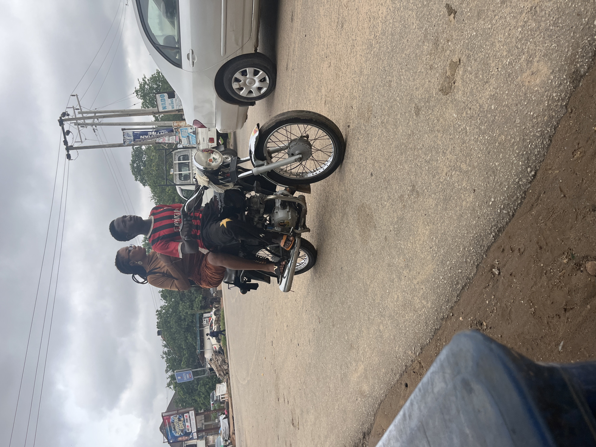 Two individuals are riding on a motorcycle on a paved road. The person in front, presumed to be the driver, is a young male wearing a red and black striped t-shirt and black pants with a yellow star emblem. The person behind is a young female wearing a brown dress and glasses. Both are looking forward. The motorcycle is a standard model with a visible engine and exhaust pipe. In the background, there are trees, utility poles with power lines, and a silver car. A billboard with advertisements is also visible on the left side of the image. The sky is overcast. The scene appears to be a street in Abuja, Nigeria, likely a commercial or residential area given the presence of shops and infrastructure.