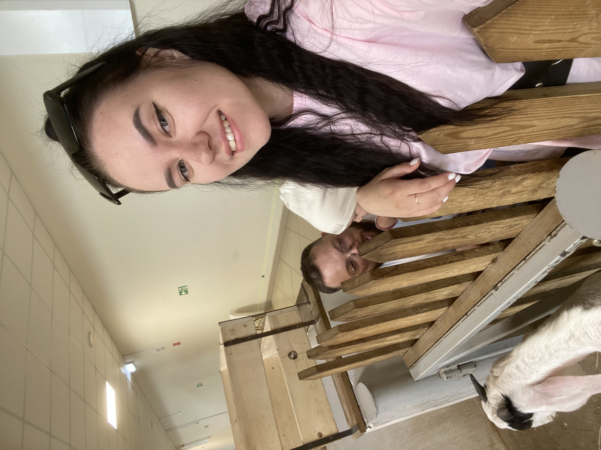 A rotated, high-angle selfie shows a woman with long, dark curly hair and sunglasses perched on her head, smiling at the camera. She wears a pink shirt and a black belt. A man with a beard is visible behind her, also smiling. They both lean over a wooden structure of horizontal slats, possibly a fence. A small goat with white fur and black markings is partially visible in the lower right corner, near the wooden structure. The background is a plain, brightly lit interior with tiled walls and ceiling. A sign with a green arrow is on the ceiling. The location appears to be indoors at a farm or petting zoo. The photo was potentially taken in the city of Perm, Russia.
