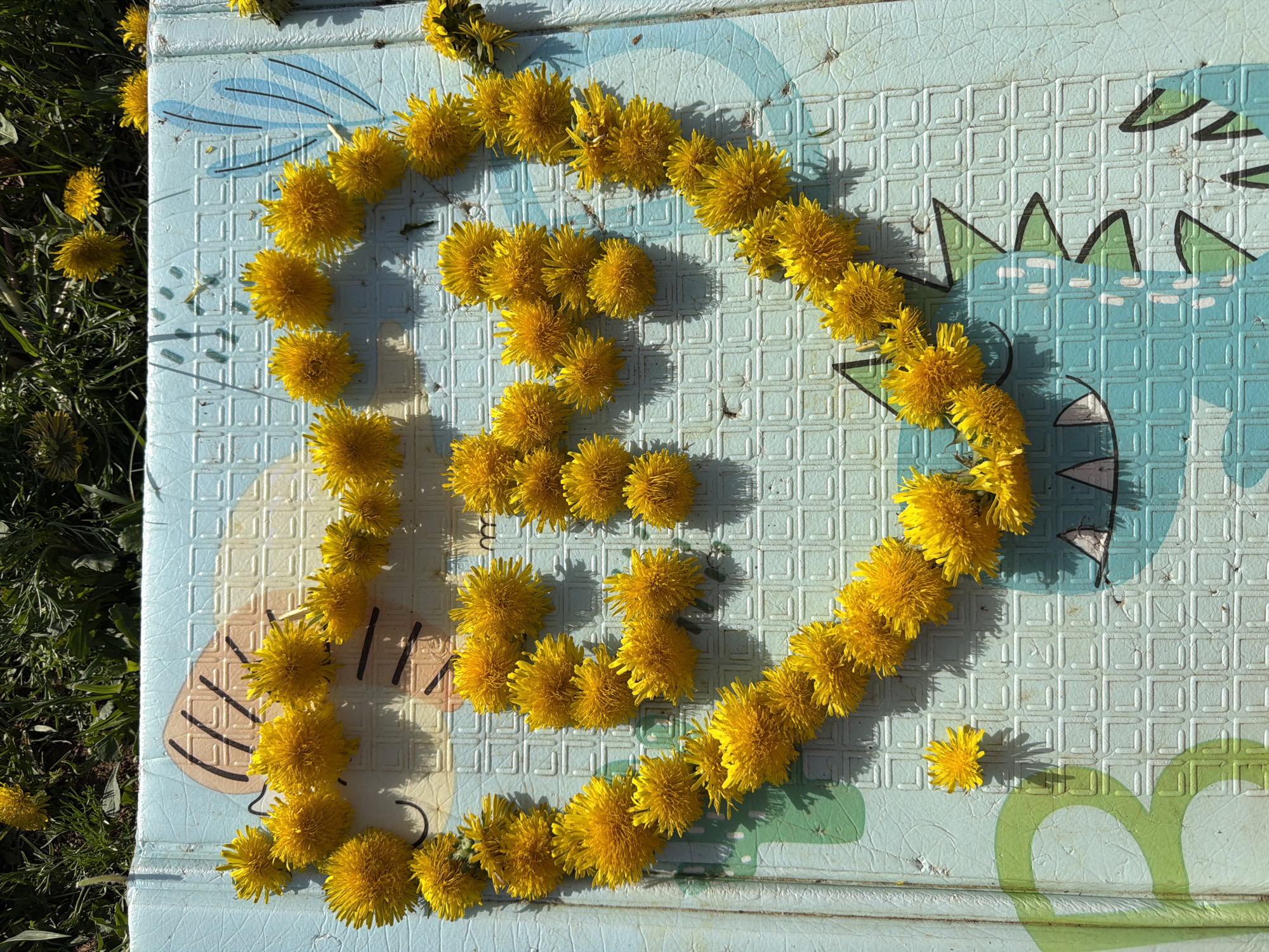 The image depicts a blue and white patterned mat, likely an outdoor play mat, partially positioned on grass. The mat has a dinosaur design on the right side. Arranged on the mat are yellow dandelions, forming the shape of a letter or symbol. Some dandelions are scattered near the grass edge and elsewhere on the mat. The location is assumed to be Chaikovsky, Russia. The image composition suggests a natural setting. There are no apparent people present in the image.