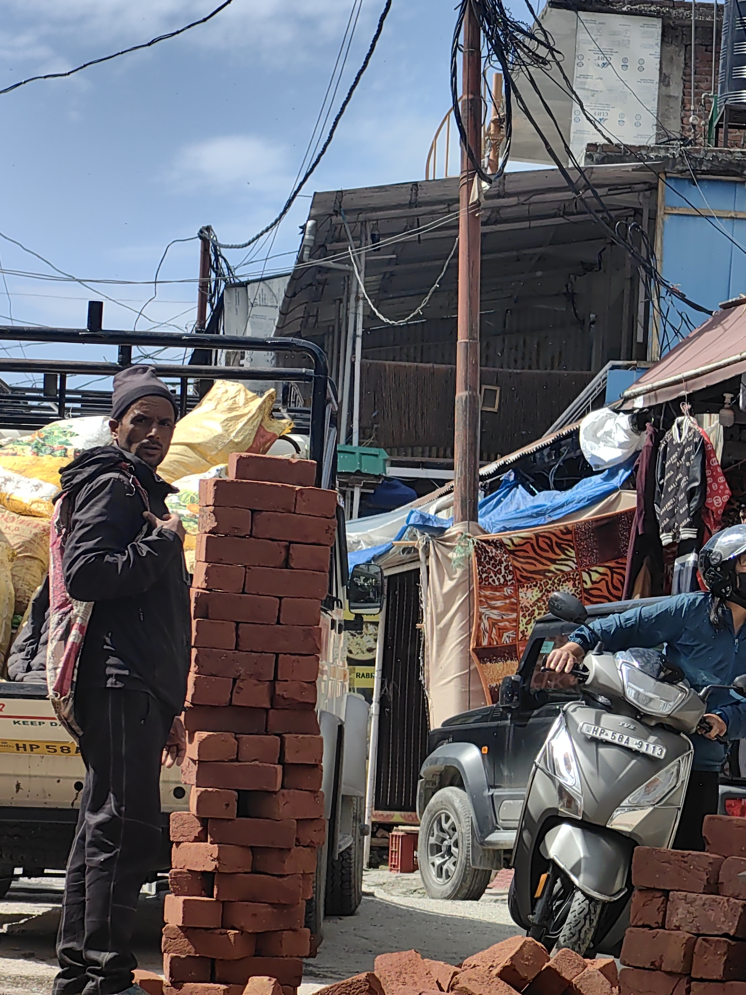 This image captures a street scene in Vashisht, India. A man in a black jacket and cap stands beside a stack of red bricks, looking towards the camera. He has a bag slung over his shoulder. In the background, a truck is loaded with yellow sacks, and a dark-colored SUV is parked. A man on a grey scooter, wearing a blue jacket and helmet, is also visible. The scene is cluttered with electrical wires and corrugated metal roofing, suggesting an urban or semi-urban environment.