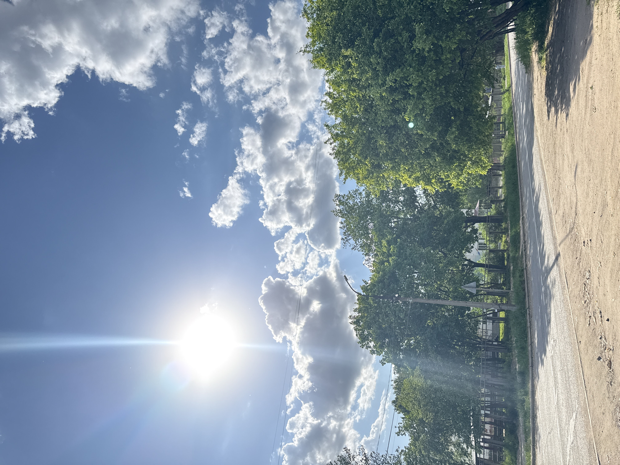 The image depicts a view of the sky, trees, and a road. The sky is blue with scattered white clouds. The sun is visible, creating a lens flare. On the right side of the image, there are trees with green foliage. A road runs alongside the trees, with a strip of dirt or gravel visible on the right edge. The image appears to be taken from a ground-level perspective. It is of the city of Chaikovsky, Russia.