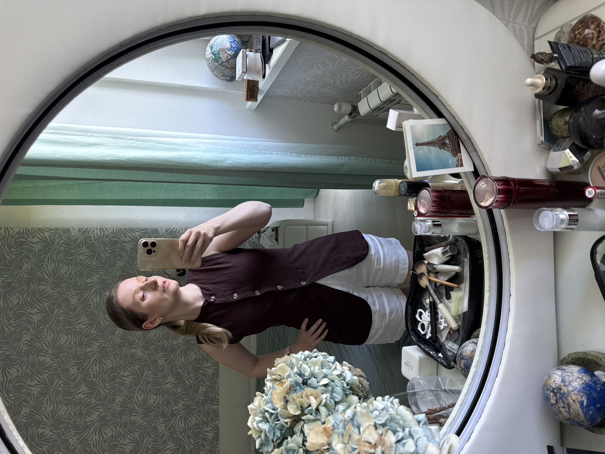 A woman stands in front of a mirror, taking a selfie. She has blonde hair pulled back in a ponytail and is wearing a sleeveless maroon vest and white shorts. The mirror reflects a bedroom with patterned wallpaper and a bed in the background, as well as a vanity with various cosmetic items and a globe. A bouquet of dried hydrangeas is visible on the left side of the frame. The lighting suggests it is daytime.