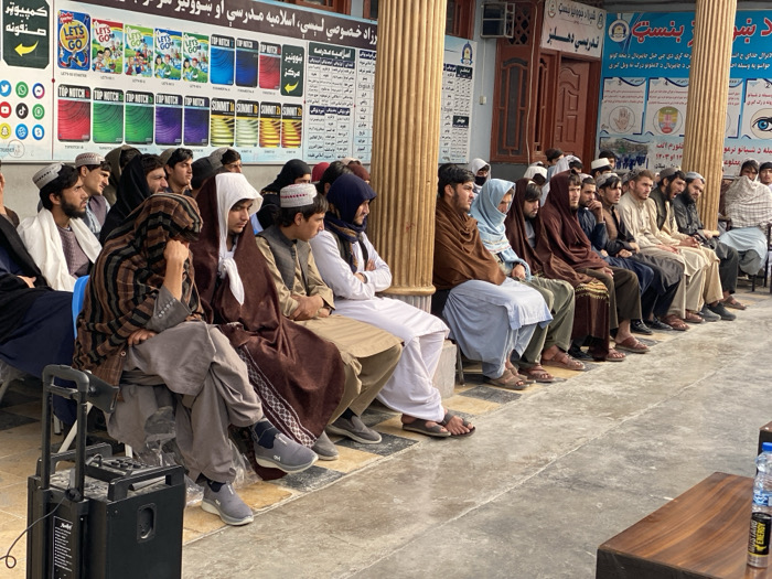 The image depicts approximately 20-25 Afghan males, ranging from young adults to middle-aged, seated in rows on blue plastic chairs within an outdoor or semi-enclosed area of the Benast Educational Center in Kandahar, Afghanistan. They are dressed in traditional shalwar kameez, vests, and various head coverings including pakols, turbans, and shawls draped over their heads or shoulders. Their attention is directed towards the right, out of frame, suggesting they are observing or listening to a speaker or event. One man in the immediate foreground on the left sits with his chin resting on his hand.
The background consists of a building with cream-colored walls, reddish-brown wooden doors, and decorative pillars. A large white sign mounted on the wall displays text in Dari/Pashto, covers of "Let's Go," "Top Notch," and "Summit" English language textbooks, and social media icons (Facebook, Twitter, YouTube, TikTok, Instagram, WhatsApp, Telegram). Another sign clearly identifies the institution as "بنست ښوونیز مرکز" (Benast Educational Center).
In the left foreground, a black portable speaker unit with wheels is visible. On the right, a segment of a wooden table or podium is visible, holding a plastic water bottle and a yellow Monster Energy drink can. The ground is paved with concrete, displaying faint square patterns.