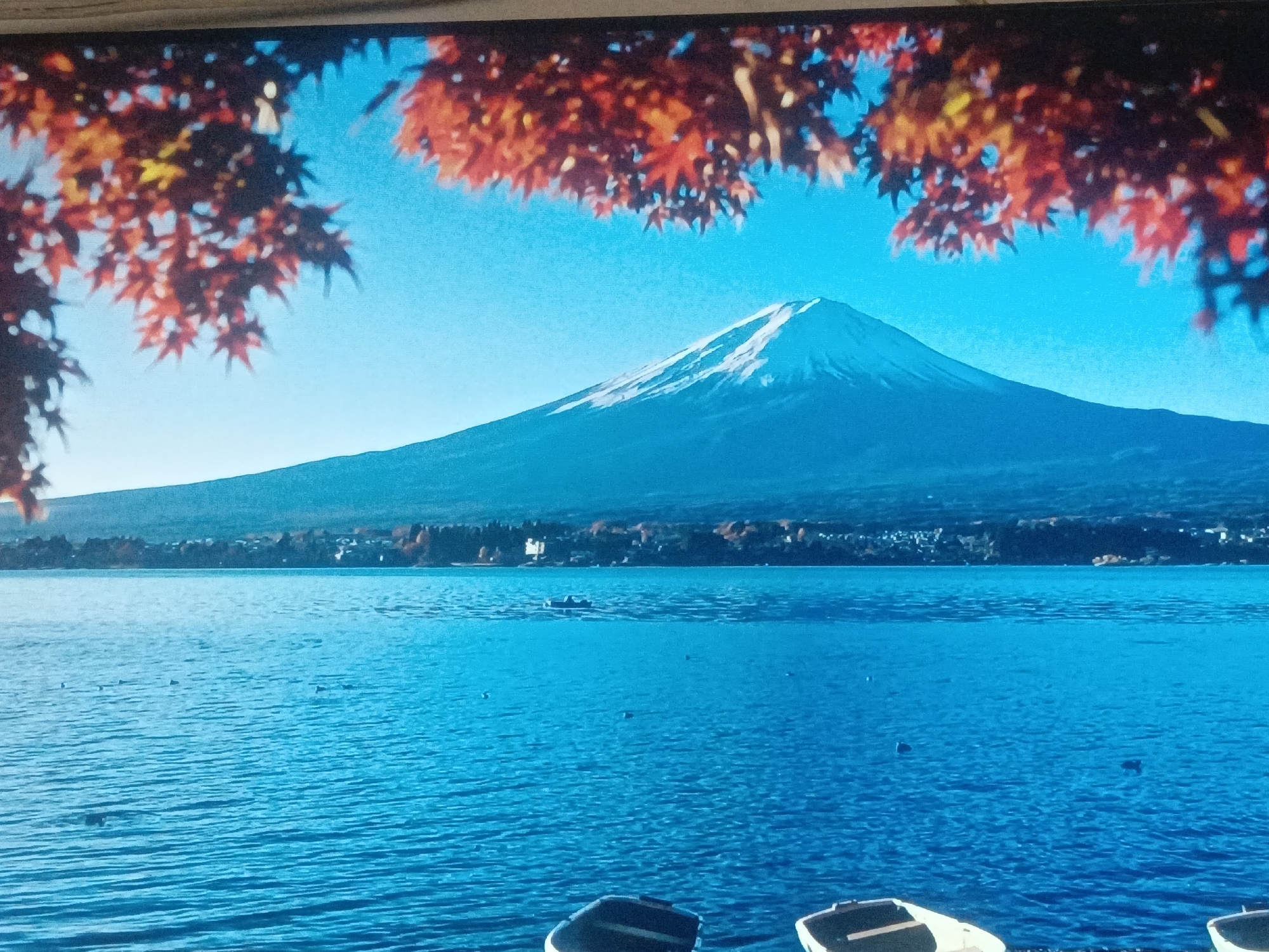 The image depicts a serene landscape featuring Mount Fuji in Japan. The mountain is centrally positioned, its peak capped with snow, set against a clear blue sky. The foreground showcases a large body of water, likely a lake, with gentle ripples on its surface. Several small boats are visible near the bottom of the frame. The upper portion of the image is framed by autumnal foliage with red leaves. The location is not in Juan Griego, Venezuela.
