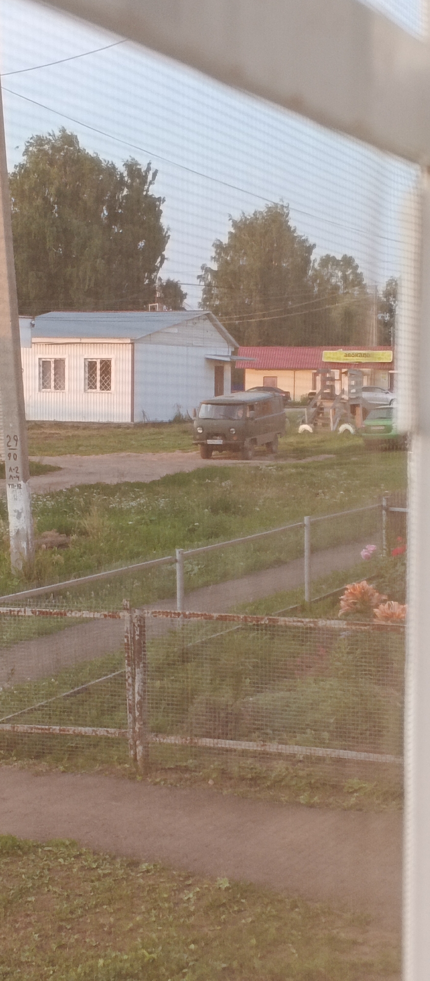 The image depicts a view through a screen in the village of Podgornoye, Russia. In the foreground, a gravel path is visible, bordered by a rusty wire fence. Beyond the fence, a light-colored building with two windows is situated. To the right of this building, a green van is parked. Further in the background, there are trees and another building with a red roof. The overall scene suggests a rural or semi-rural environment.