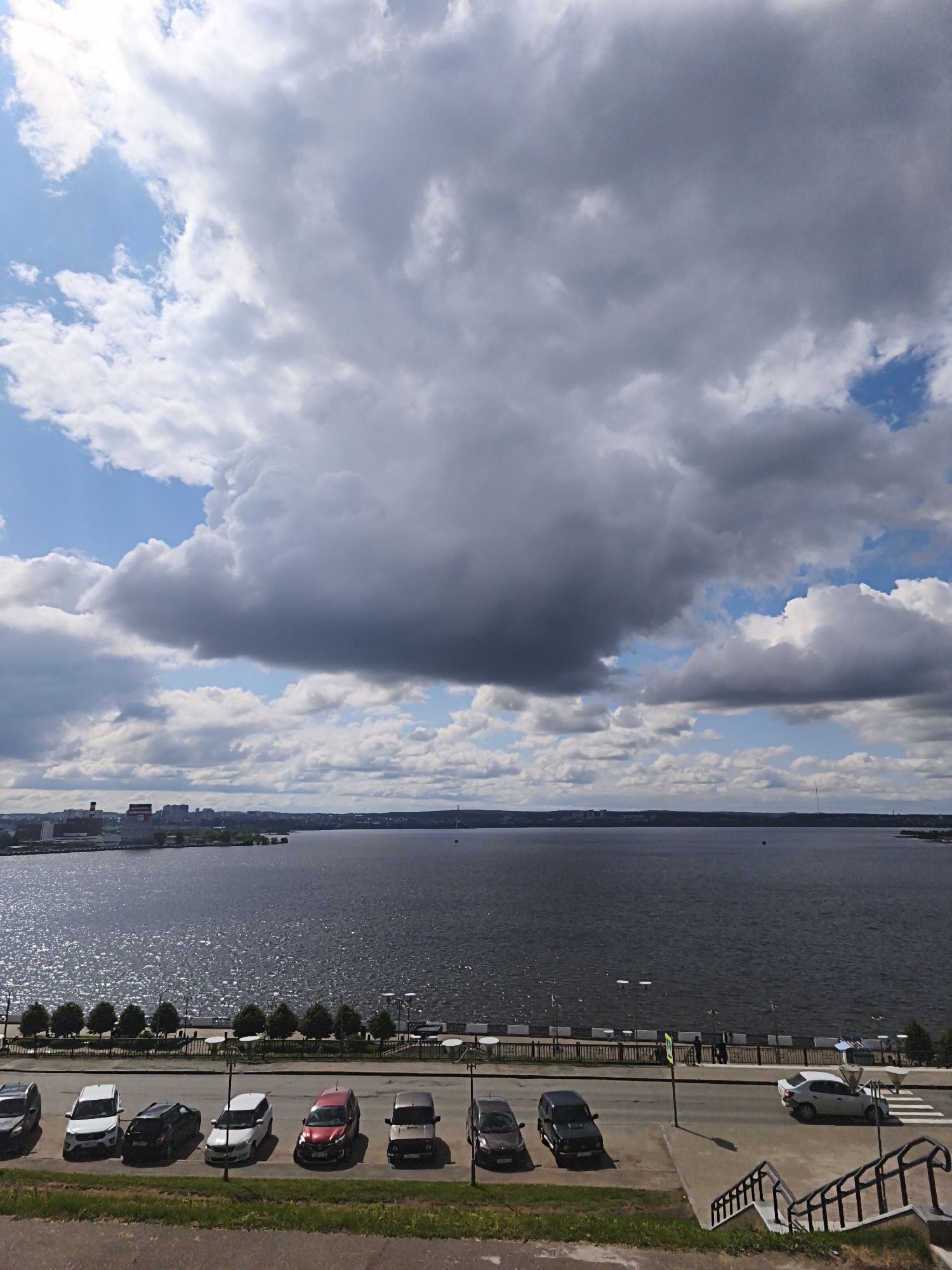 This image presents a wide, elevated view of a large body of water, likely a river or lake, with a city skyline visible on the distant bank under a dramatic sky.
The sky is dominated by vast, heavy cumulus clouds, predominantly dark grey and suggesting an overcast or changeable weather condition, though patches of brighter blue sky with smaller white clouds are visible, especially towards the left. Sunlight glints off parts of the water's surface, indicating it is daytime.
Across the water, the silhouette of a city with various buildings, including what appears to be industrial structures and taller residential or commercial blocks, stretches along the horizon. On the near bank, a paved promenade lines the water's edge, featuring a row of evenly spaced small trees, streetlights, and a protective fence. A few individuals can be seen strolling along this promenade.
In the foreground, a paved road runs parallel to the waterfront. A line of cars is parked along the side of this road, including a dark grey SUV, a white SUV, a black sedan, a red hatchback, another dark grey SUV, and a beige/silver SUV. Further down and to the right, another section of road shows a white car near a crosswalk, and a dark brown car and a black car parked. A set of metal stairs and railings leads downwards in the bottom right corner.
Visible text includes partial license plate numbers on some of the parked vehicles, such as "18" on a white SUV and "16" and "116" on other cars, which are regional codes for Udmurtia and Tatarstan in Russia, respectively, confirming the geographic context of Izhevsk, Russia. The overall scene depicts a typical cloudy day at a city waterfront, with people going about their daily routines.