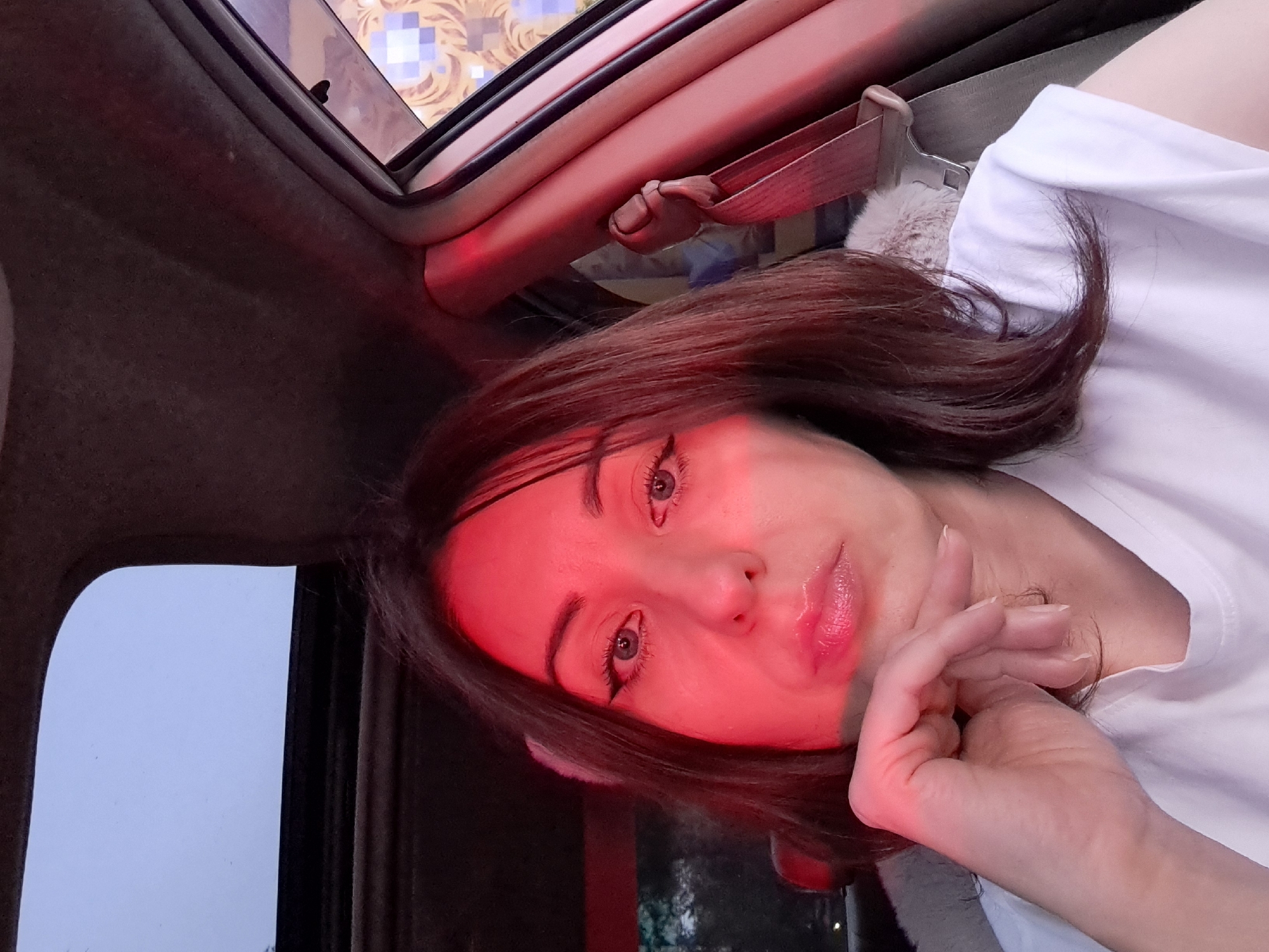 A woman with brown hair is sitting in the passenger seat of a car. She is looking at the camera and resting her chin on her hand. She is wearing a white t-shirt. The car has a sunroof. The lighting is dim and red. The background is blurred. There is a seatbelt visible. The image does not provide enough information to determine the location in the city of Irkutsk, Russia.