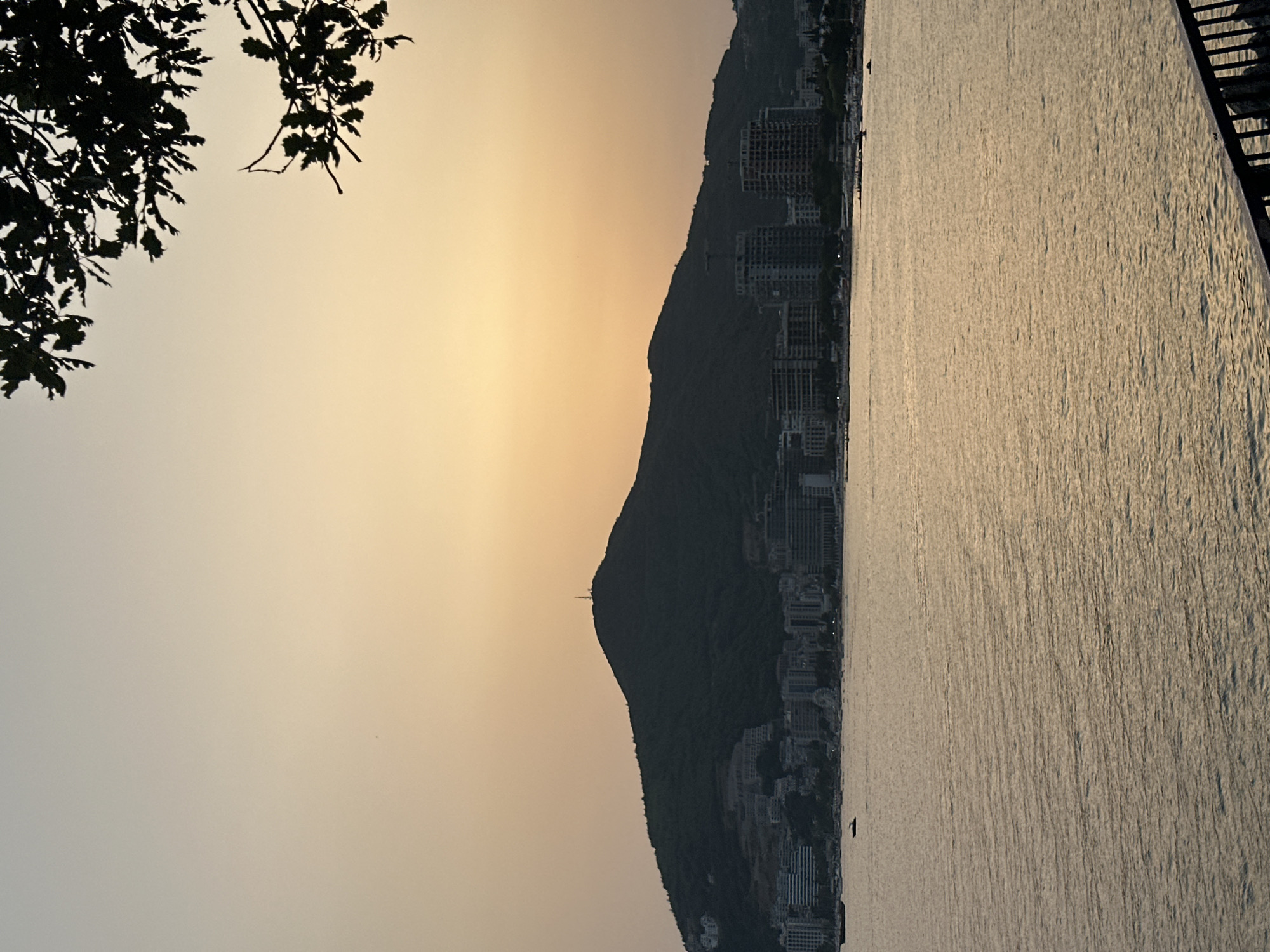 The image shows a sunset over the Adriatic Sea with the city of Budva, Montenegro, in the background. A large, dark green mountain dominates the center of the frame, with a telecommunications tower visible at its peak. The city is nestled at the base of the mountain, with rows of buildings and construction sites. The sea is calm, with a few small boats visible. The sky is a gradient of orange and yellow, with tree branches silhouetted in the upper right corner. The lighting suggests it is dusk.