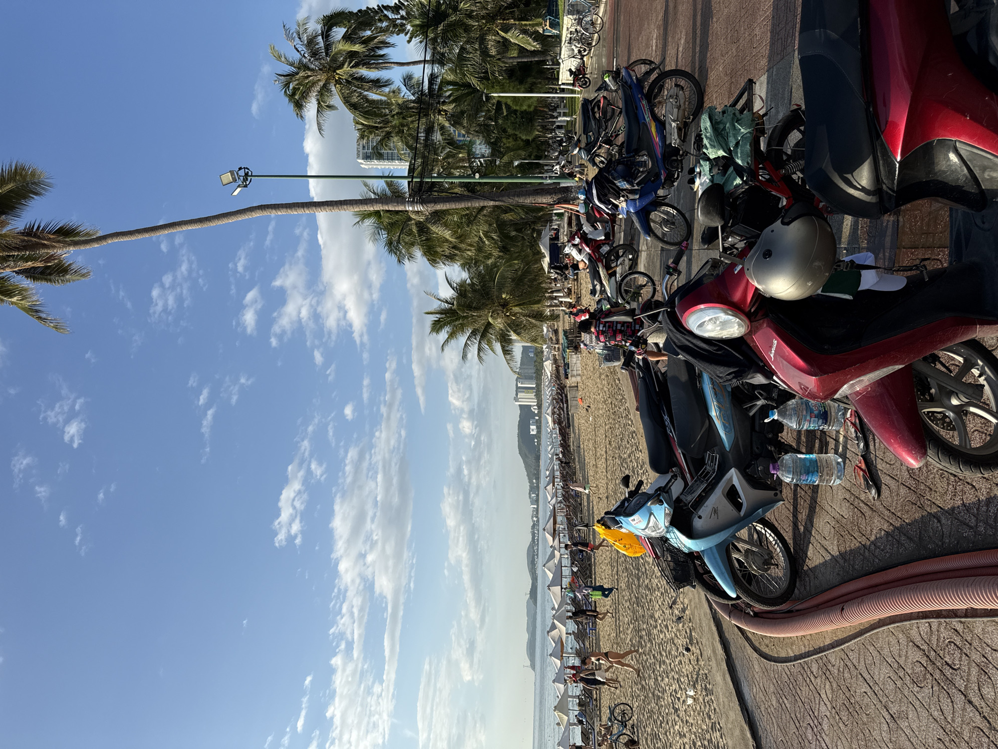 This image captures a sunny day at a beach in Nha Trang, Vietnam. The foreground is dominated by several parked scooters and motorcycles, with a red one on the right and a blue one to its left. Two bottles of water and a pair of flip-flops are on the ground near the blue scooter. Behind the scooters, a sandy beach stretches out, lined with rows of white beach umbrellas and loungers. People are visible on the beach, some walking, some sitting, and others in the shallow water. Further back, palm trees line the promenade, and taller buildings are visible in the distance, characteristic of Nha Trang's coastal cityscape. The sky is blue with scattered white clouds, and a tall lamppost stands prominently near a palm tree. The overall scene depicts a vibrant beachside environment with typical urban coastal elements.