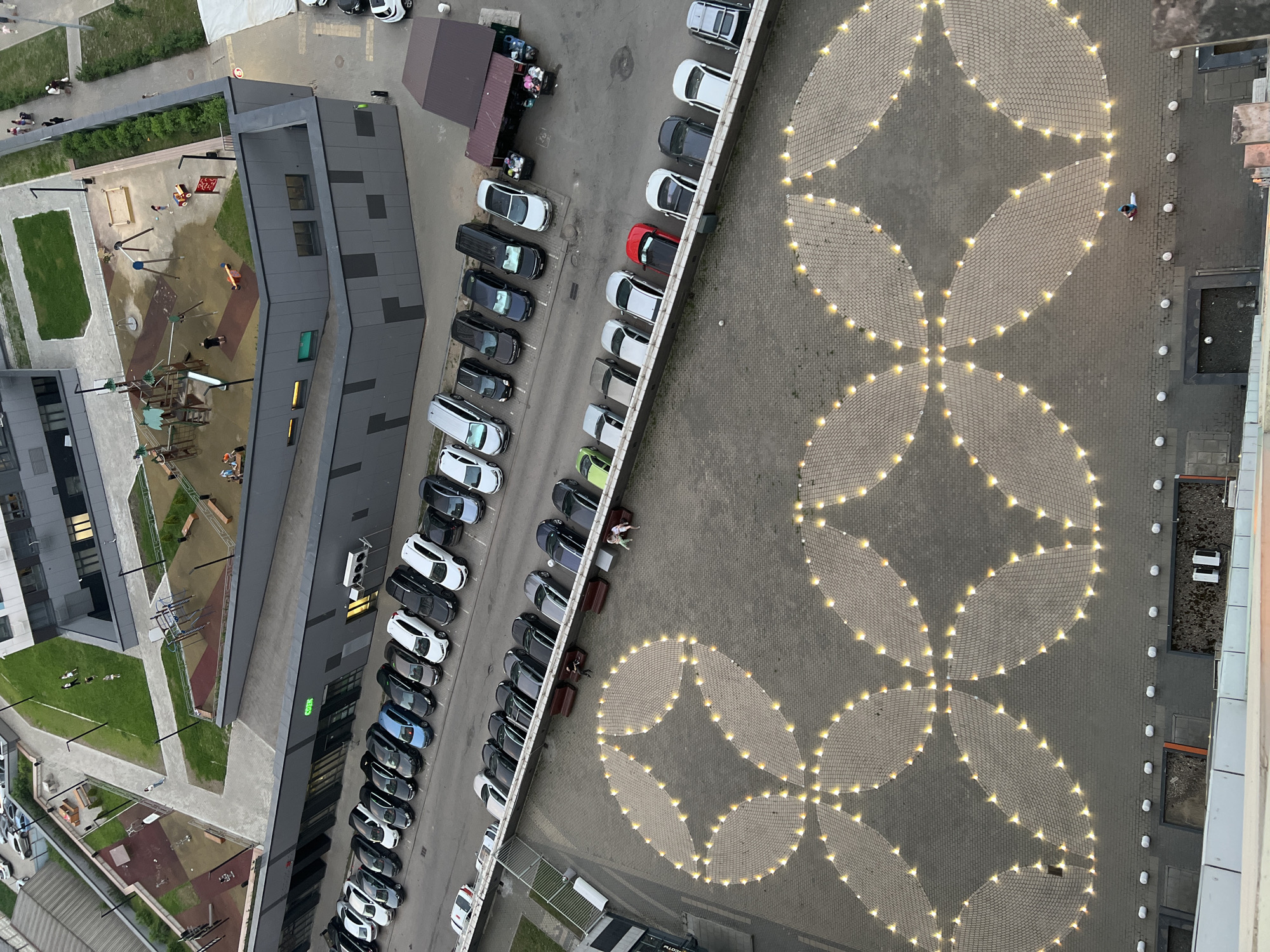 An aerial shot shows a residential area in Mytishchi, Russia. A playground with wooden structures and a sandbox is on the left. A modern building with a gray facade stands next to the playground. Adjacent to the building, a parking lot is filled with various cars. A pedestrian area decorated with patterns of interconnected circles formed by lights is visible on the right. People are sitting on benches in the pedestrian area.