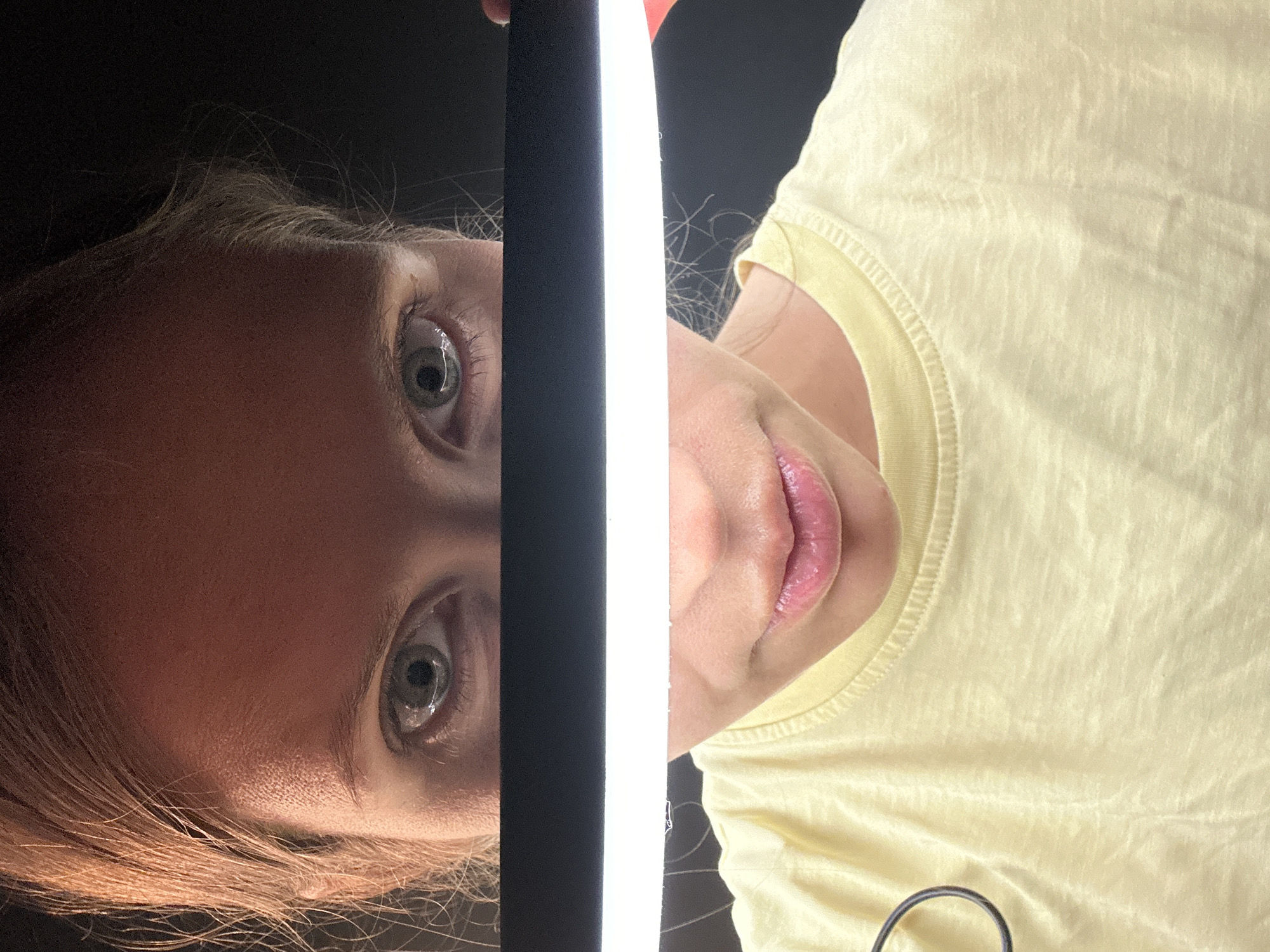 A close-up photograph displays half a person's face on the left, defined by an eye with a visible tear and red hair. A light source is positioned vertically in the center of the frame. On the right, the individual's lips and the upper portion of a yellow t-shirt are visible. The background behind the face and shirt is black. The location in Fethiye, Turkey, is not determinable from this image.