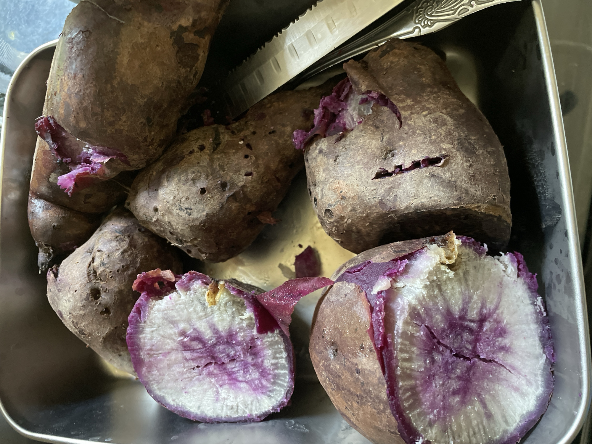 A close-up, top-down shot captures a collection of purple sweet potatoes in a metallic tray. Some of the sweet potatoes are whole, while others have been cut open, revealing their vibrant purple and white flesh. A silver knife with a serrated edge rests beside some of the potatoes. The lighting is natural, casting subtle shadows and highlighting the textures of the sweet potatoes and the tray. The setting appears to be a kitchen counter. The image does not provide information about its location in Davao City, Philippines.
