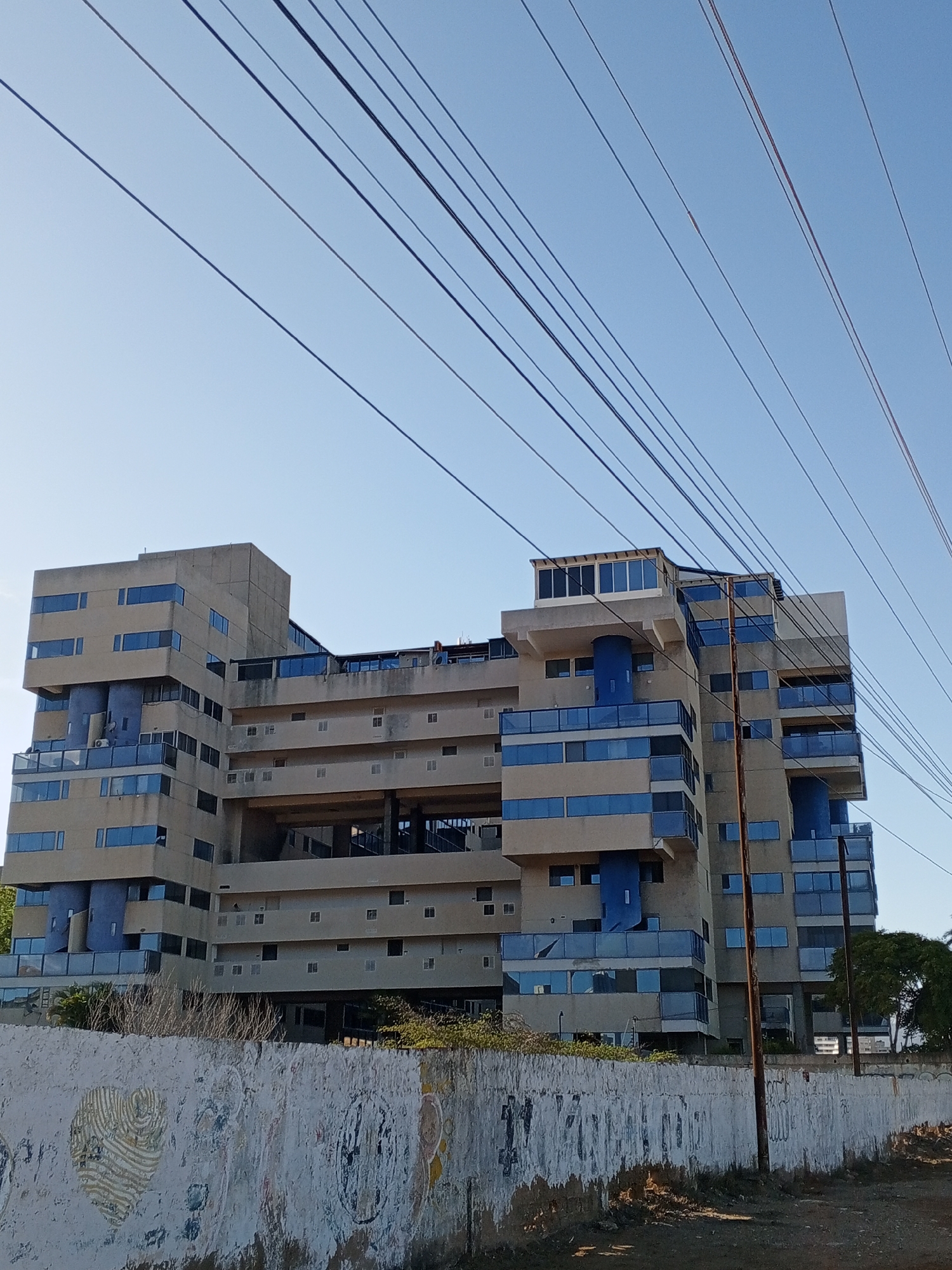 The image captures a static, daytime scene featuring a large, multi-story building under a clear blue sky. Dominating the upper portion of the frame are numerous dark power lines, stretching diagonally across the sky from left to right.
The central subject is a substantial building with a unique, stepped, and modular design. Its facade is primarily composed of light brown/beige concrete, contrasting with numerous blue-tinted windows, some arranged in horizontal strips, and blue glass railings on balconies. Several sections of the building feature prominent blue cylindrical elements or wall panels. Some areas appear unfinished or exposed, revealing internal structures, particularly in the center. A distinctive glass-enclosed structure sits on the rooftop of one section. The architecture suggests a modern, possibly Brutalist influence, indicating a large apartment complex or hotel.
In the foreground, a weathered and textured concrete wall spans the width of the image. It is covered in peeling white paint, revealing patches of the underlying surface, and features faded, abstract artistic designs, including a stylized heart shape on the left. The ground in front of the wall is bare and dusty, with some sparse, dry vegetation visible behind it. A utility pole stands on the right side of the frame, extending vertically towards the building.
The bright, clear sky suggests a sunny day, likely morning or late afternoon, given the quality of light. There is no human activity visible in the scene, and no discernible text is legible on any object. The overall impression is one of an urban or semi-urban environment in Porlamar, Venezuela, with a blend of structured architecture and aged street elements.