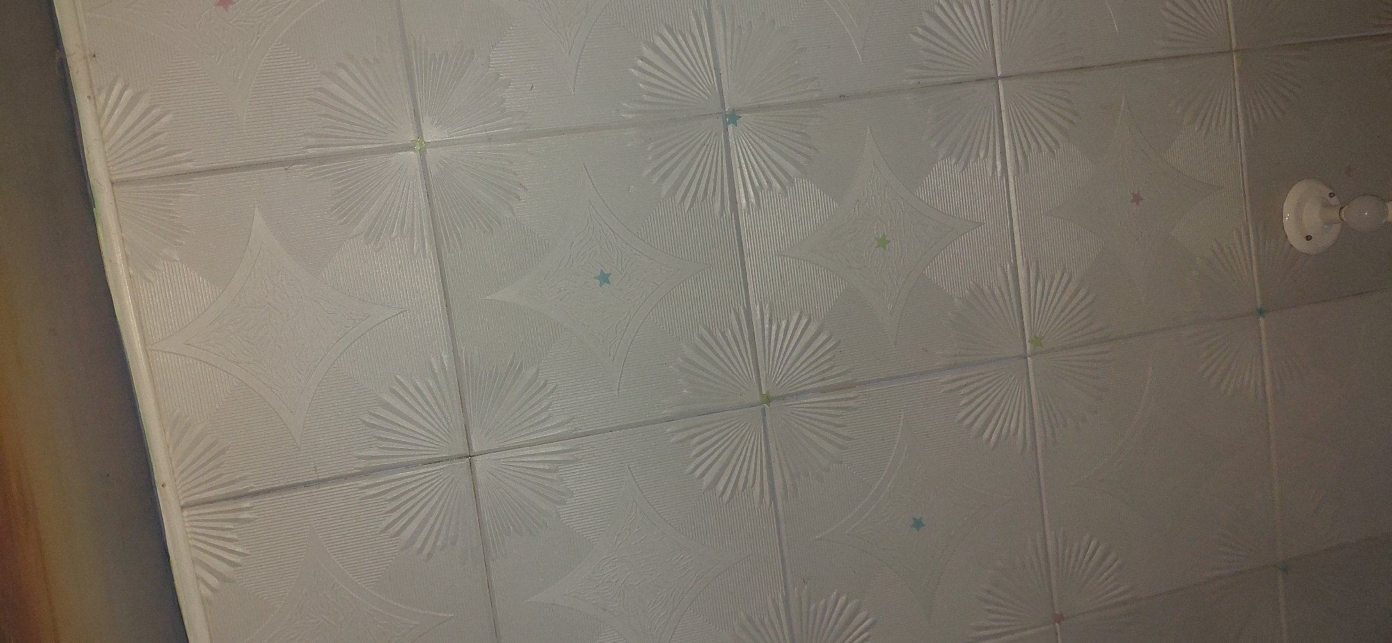 The image shows a white patterned ceiling made of square tiles. The tiles have a repeating design of radiating lines and star-like shapes. There are small, colored star accents scattered across the ceiling. In the bottom right corner of the image, a portion of a white ceiling light fixture is visible. The overall scene appears to be an interior room. There is no indication of the city of Reni, Ukraine, or any specific context that would place it there.