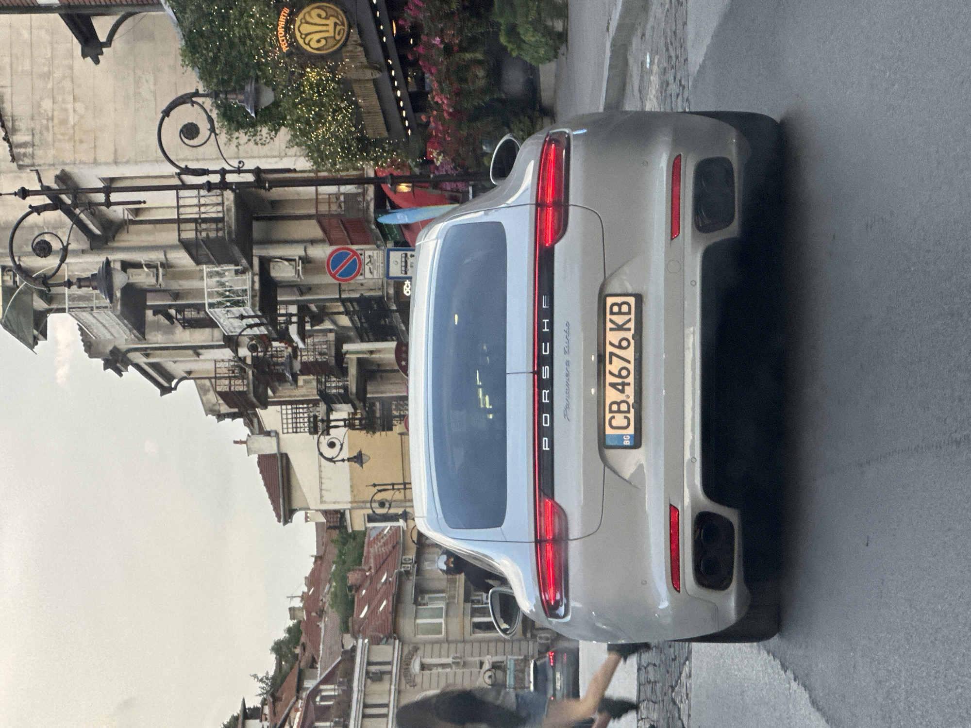 The image shows the rear of a silver Porsche Panamera parked on a street in Veliko Tarnovo, Bulgaria. The car has the license plate "CB 4676 KB". In the background, there are buildings with traditional architecture, balconies, and street lamps. A pedestrian is walking on the left side of the road, and other cars are visible in the distance. The sky is overcast.