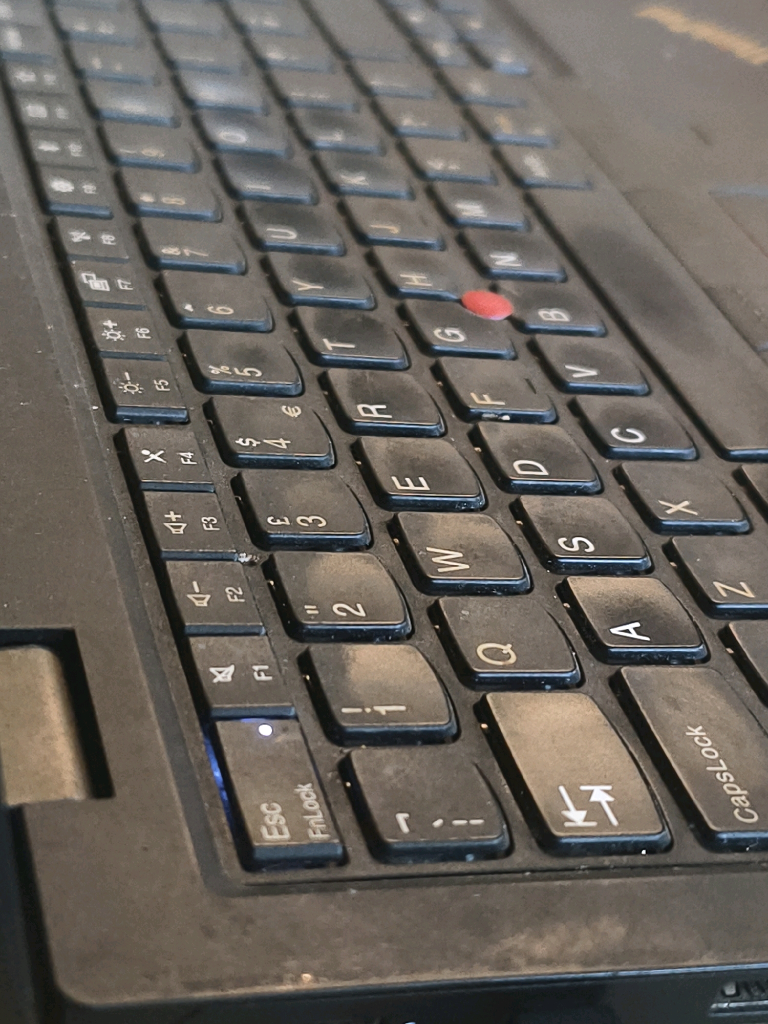 The image is a close-up, slightly angled shot of a black laptop keyboard. Dust is visible on the keys, particularly around the edges and in the crevices. The red TrackPoint in the center of the keyboard is prominent. Visible keys include the function keys (F1 through F7 with symbols and labels), alphanumeric keys (including Q, W, E, R, T, Y, U, I, O, P, A, S, D, F, G, H, J, K, L, Z, X, C, V, B, N, M), numerical keys (1, 2, 3, 4, 5), symbols ($, €, ", comma, period), and modifier keys (Esc, FnLock, CapsLock, and a key with an upward and downward arrow symbol). A blue indicator light is visible near the Esc key, and a white LED is on above it. The surrounding environment is dark and out of focus, suggesting the photo was taken in low light conditions. There is no information in the image to indicate a location in Muara Bulian, Indonesia.