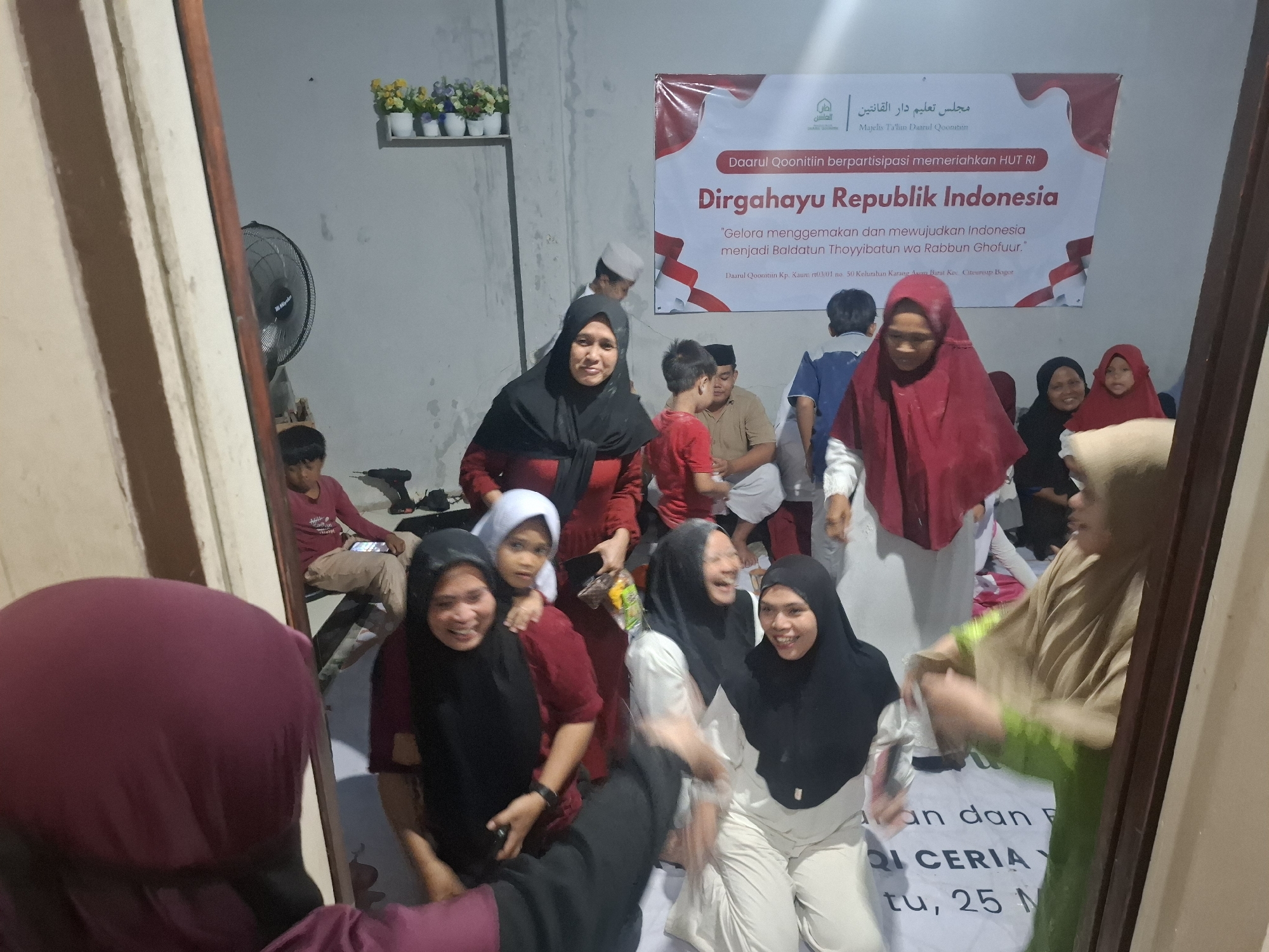 The image depicts a group of people, mostly women and children, gathered indoors. A large banner in the background, which appears to be in Indonesian, announces "Dirgahayu Republik Indonesia" and mentions "Daarul Qoonitiin berpartisipasi memeriahkan HUT RI." This suggests a celebration related to Indonesia's Independence Day. The people are dressed in casual attire, many wearing hijabs. Some individuals are smiling and interacting with each other, while others are looking towards the camera. In the foreground, a large mirror reflects some of the scene, including a person wearing a maroon hijab. The environment appears to be a room with a concrete wall, a ceiling fan, and some shelves with decorative items. The presence of a banner celebrating Indonesian Independence and the text "Daarul Qoonitiin" strongly suggests the location is within or associated with this institution in Babakan Madang, Indonesia.