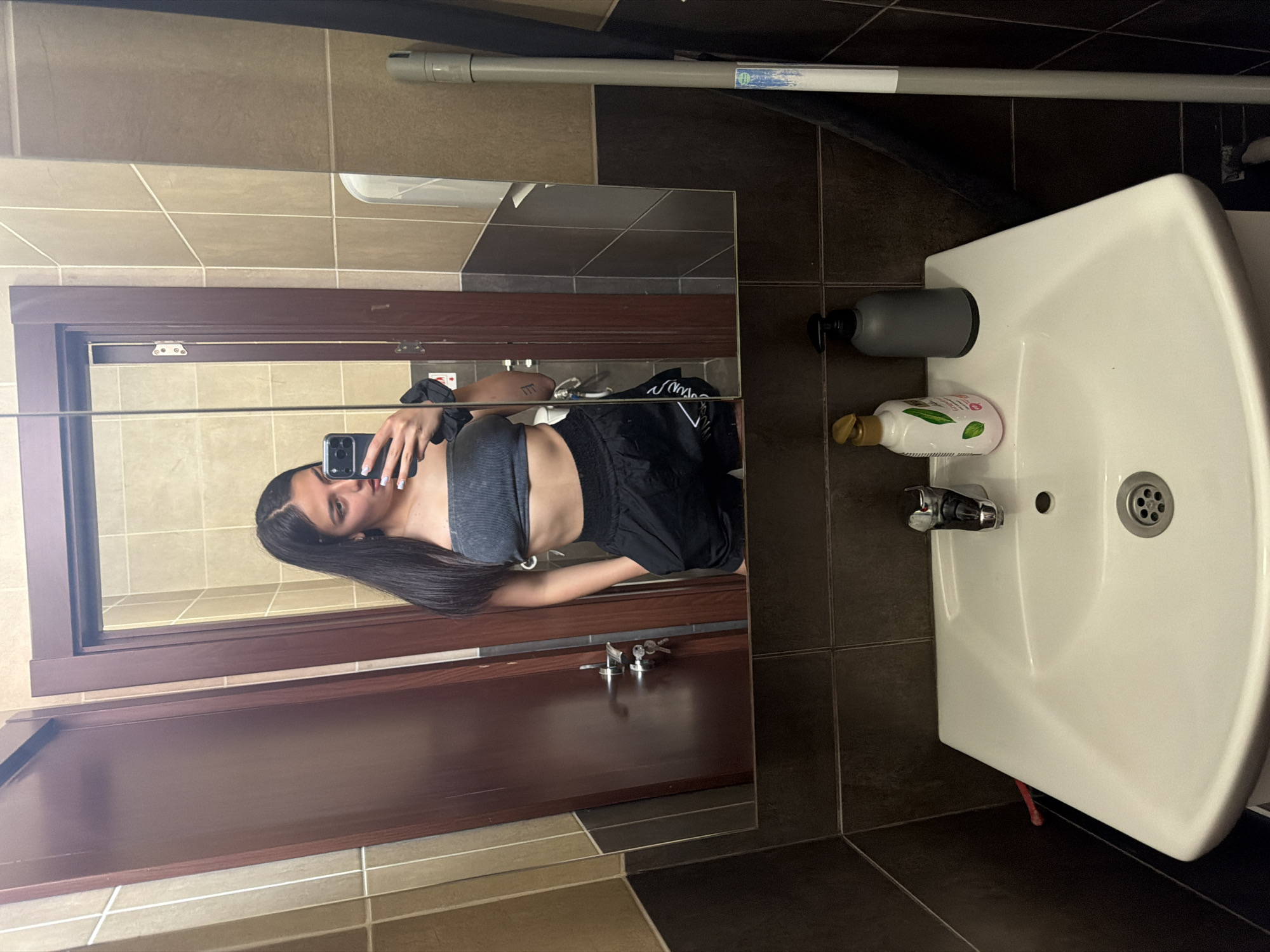 An indoor mirror selfie captures a young woman in what appears to be a bathroom, rotated 90 degrees clockwise from its natural orientation. The scene is set in Gabrovo, Bulgaria.
The subject, a young woman with long, dark hair and light-colored eyes, looks towards the camera with a neutral expression. She is wearing a grey ribbed crop top and black shorts. A black scrunchie is on her left wrist, and a small, stylized tattoo of three vertical lines with a top horizontal bar is visible on her left forearm. She holds a black smartphone, featuring multiple camera lenses, to take the mirror selfie. Her nails are painted with light-colored polish.
The setting is a bathroom with tiled walls and a tiled floor. The upper wall sections are covered in light beige tiles, while the lower sections and the area around the sink feature darker brown tiles. The floor displays a mix of beige, light brown, and dark brown tiles. A white ceramic sink with a chrome faucet and drain is prominent on the right side of the image. On the sink's edge, a grey soap dispenser and a white bottle with a gold pump are visible. The bottle's label, partially legible, suggests "Garnier Aloe Vera."
Above the mirror, grey and black plumbing pipes run across the wall, with a blue and white sticker on one pipe that contains unreadable text. The mirror itself has a dark wooden frame. Below the mirror, mounted on a dark wooden surface, are keys. A reddish plastic object or hose protrudes from under the sink. The overall lighting suggests indoor artificial illumination.
