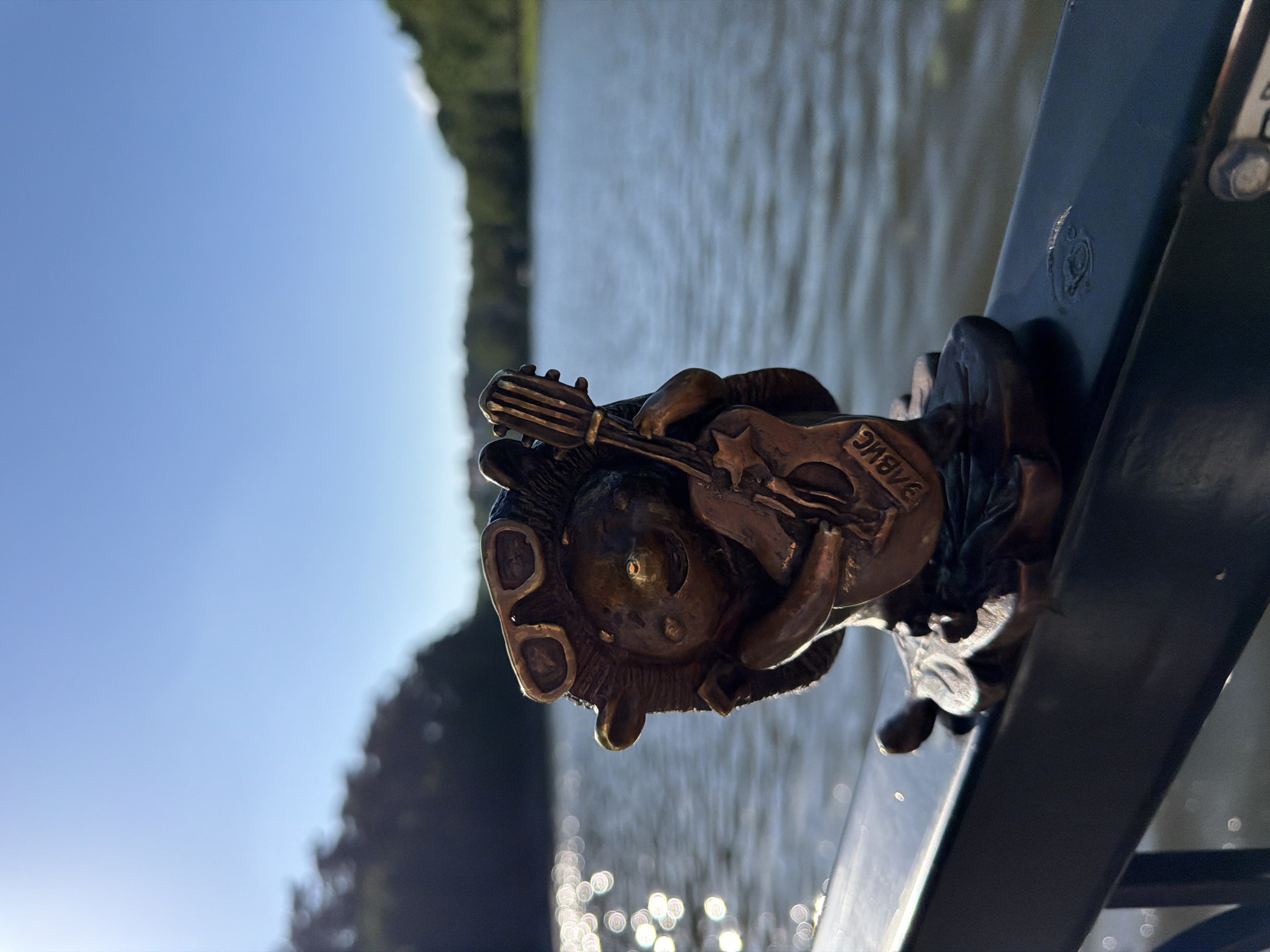 This is a close-up, horizontally oriented photograph of a small bronze figurine of a gnome playing a guitar. The figurine is attached to a dark metal railing. The gnome has a round face with large, round ears and is wearing what appear to be goggles. It is holding a miniature guitar, which has a star shape cut out of its body. Text is visible on the guitar's body, appearing to read "BABWE".
The background of the image is out of focus. To the left, a bright blue sky dominates the scene, suggesting it is daytime with clear weather. To the right of the sky, and further in the background, is a body of water, likely a lake or river, with ripples visible on its surface. Trees are visible in the upper left and right portions of the background, indicating a natural setting. The lighting suggests it is either early morning or late afternoon, given the angle of the sun and the long shadows cast. The bokeh effect from the water's surface creates circular highlights. The scene is set in Svetlogorsk, Russia.
